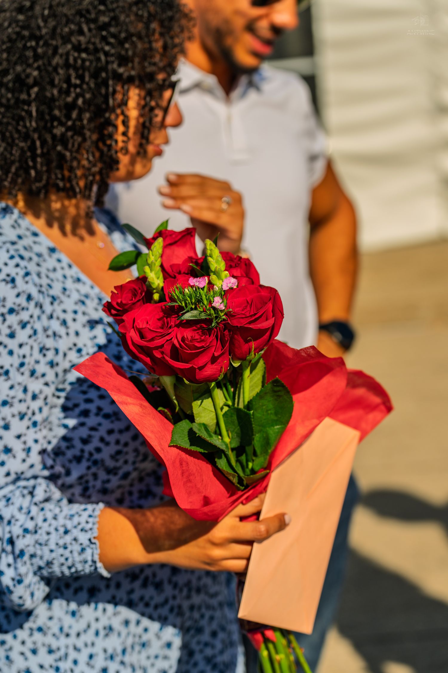 A woman with curly hair holds a vibrant bouquet of red roses while a smiling man stands behind her outdoors.