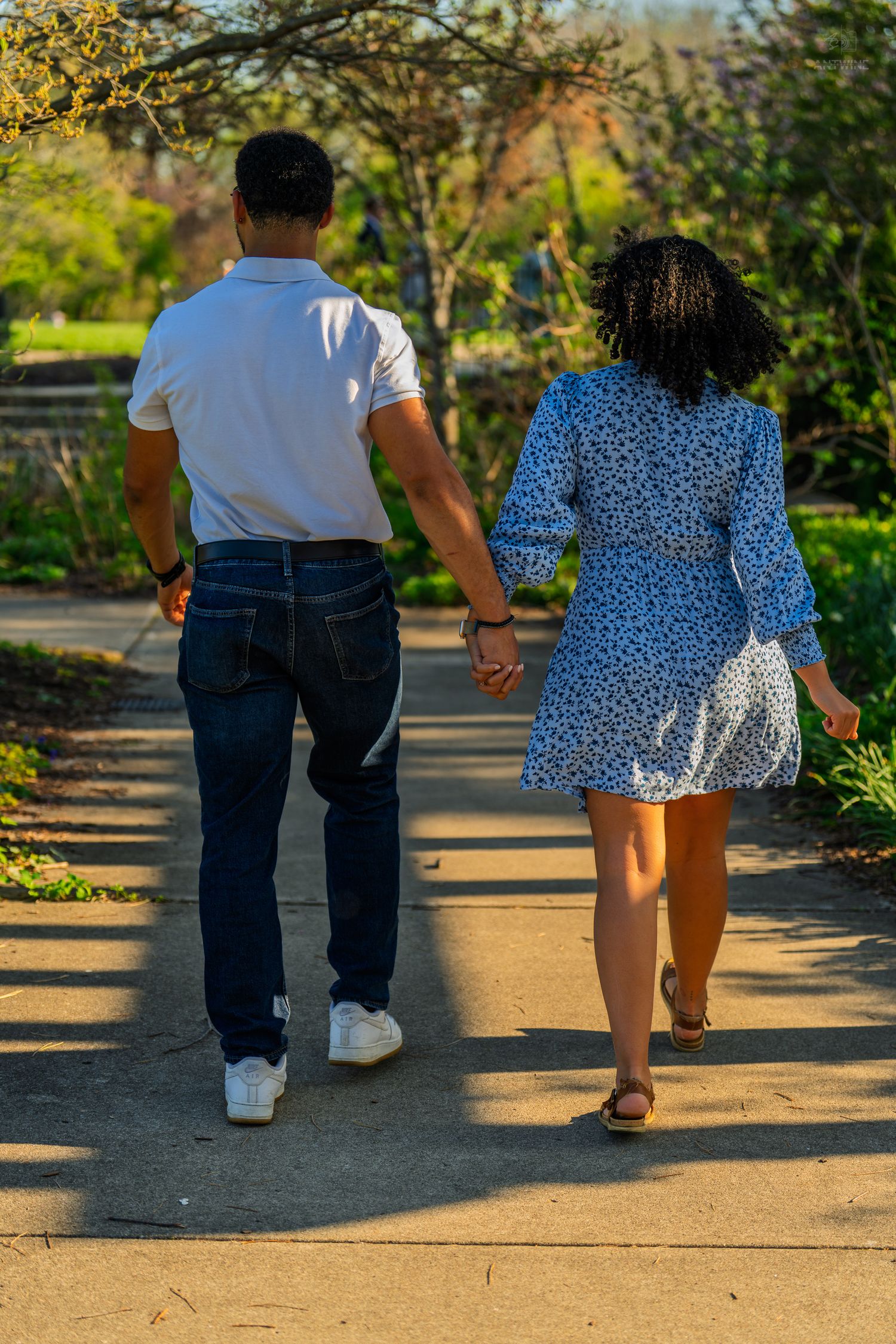 Rear view of a couple holding hands walking down a sunny park path surrounded by green foliage.