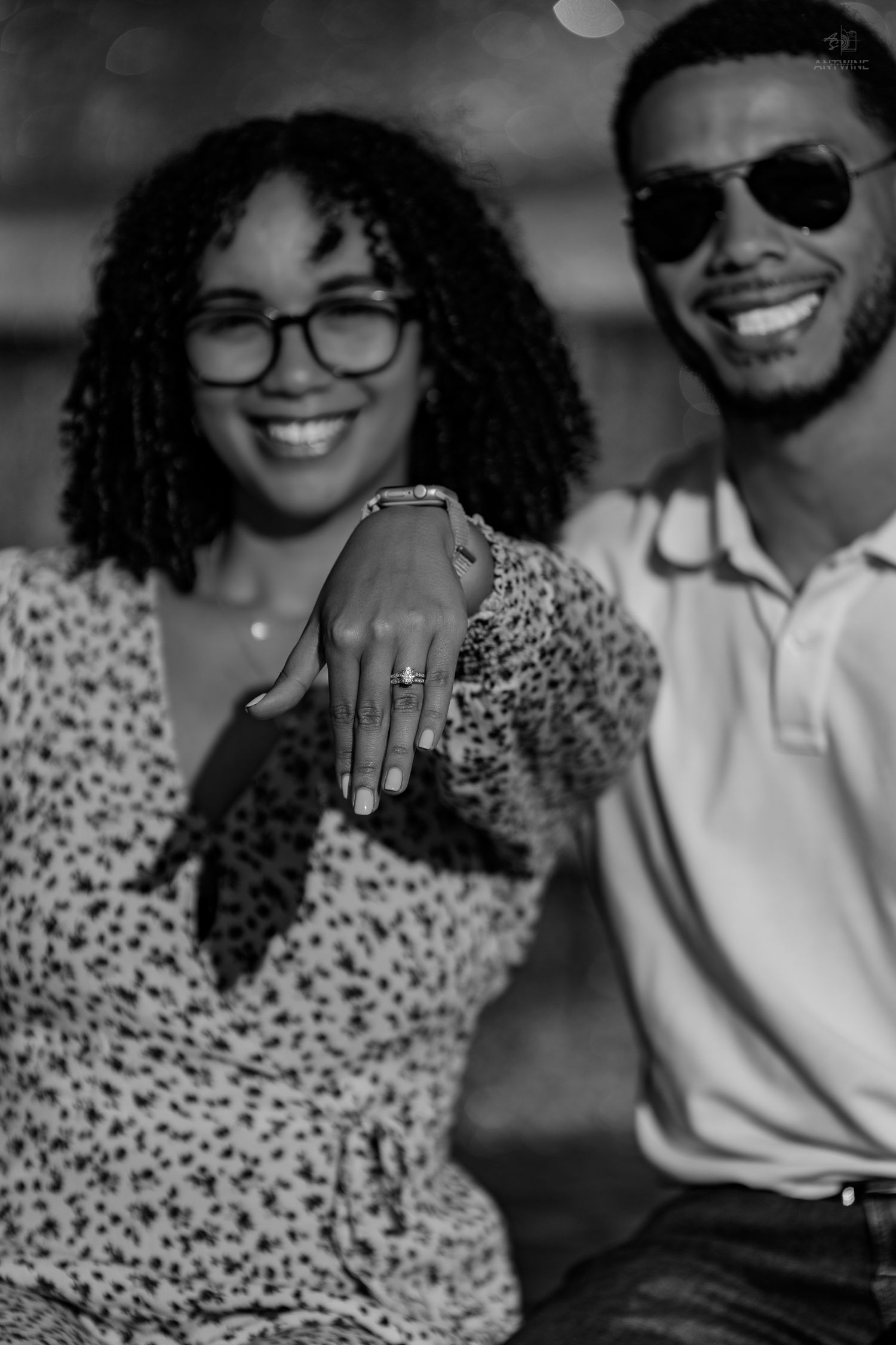 An engaged Black couple shows off an engagement ring in a black and white close-up photo.