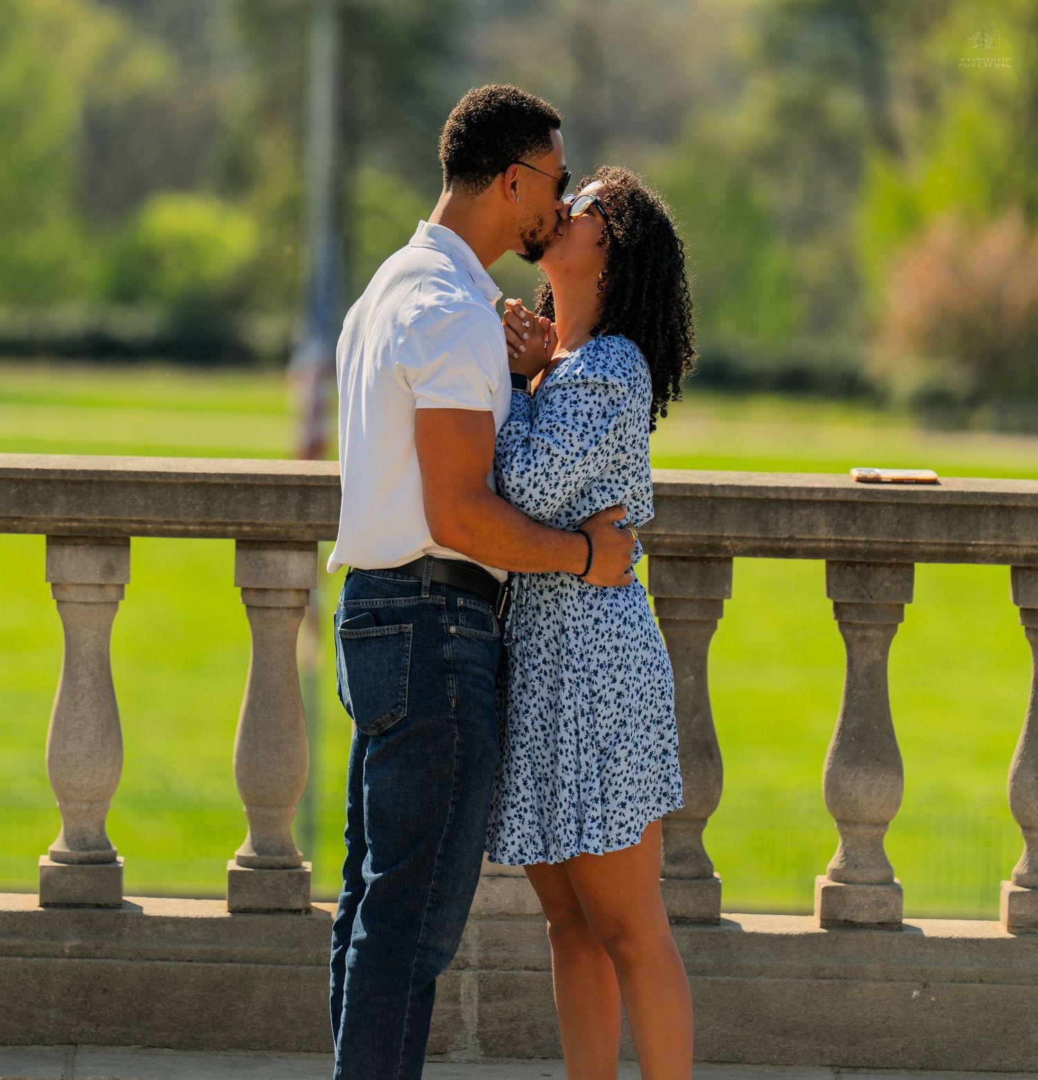 Couple sharing a kiss by decorative stone balustrade with vibrant green park grass in background.