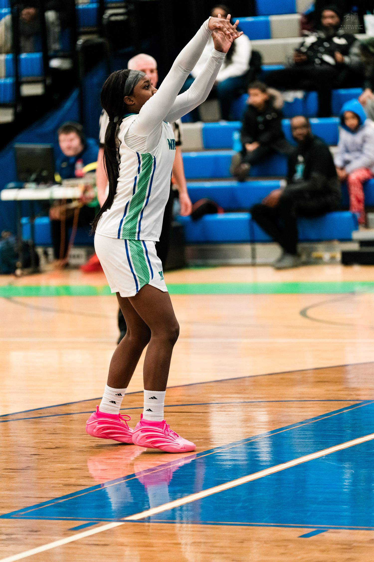 A female basketball player in a white and green Warriors uniform shoots the ball wearing bright pink sneakers indoors.