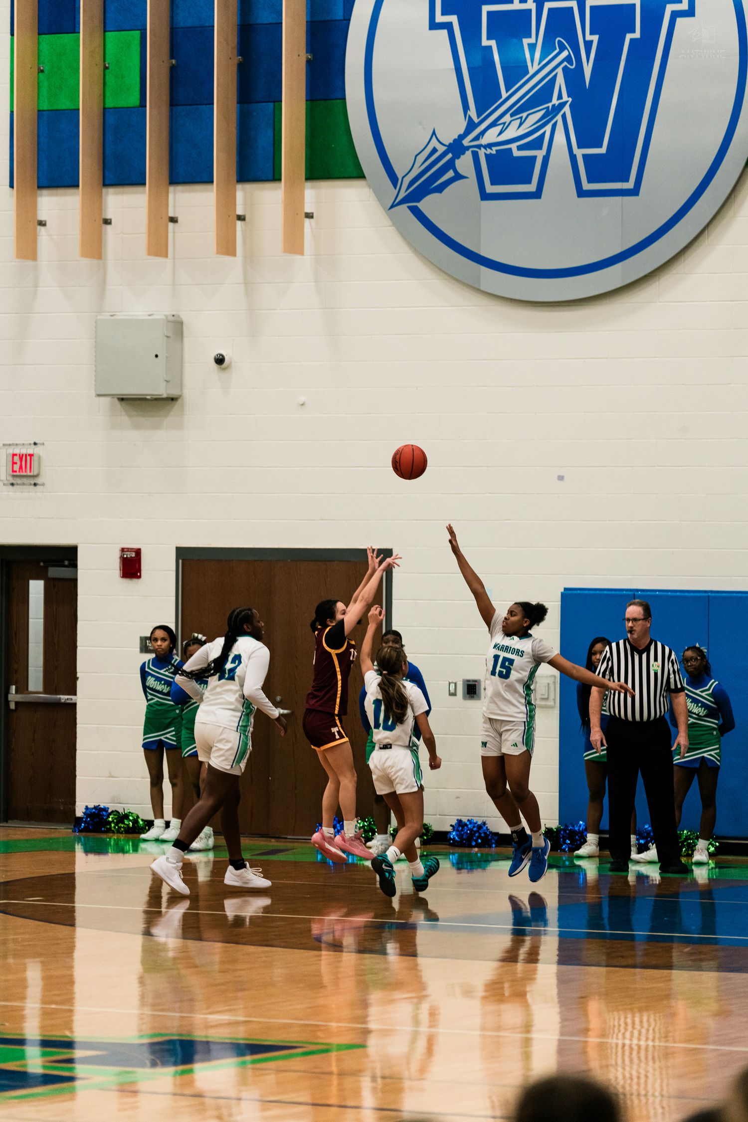 Girls basketball tip-off jump ball with Warriors players in white and teal uniforms competing in gymnasium.
