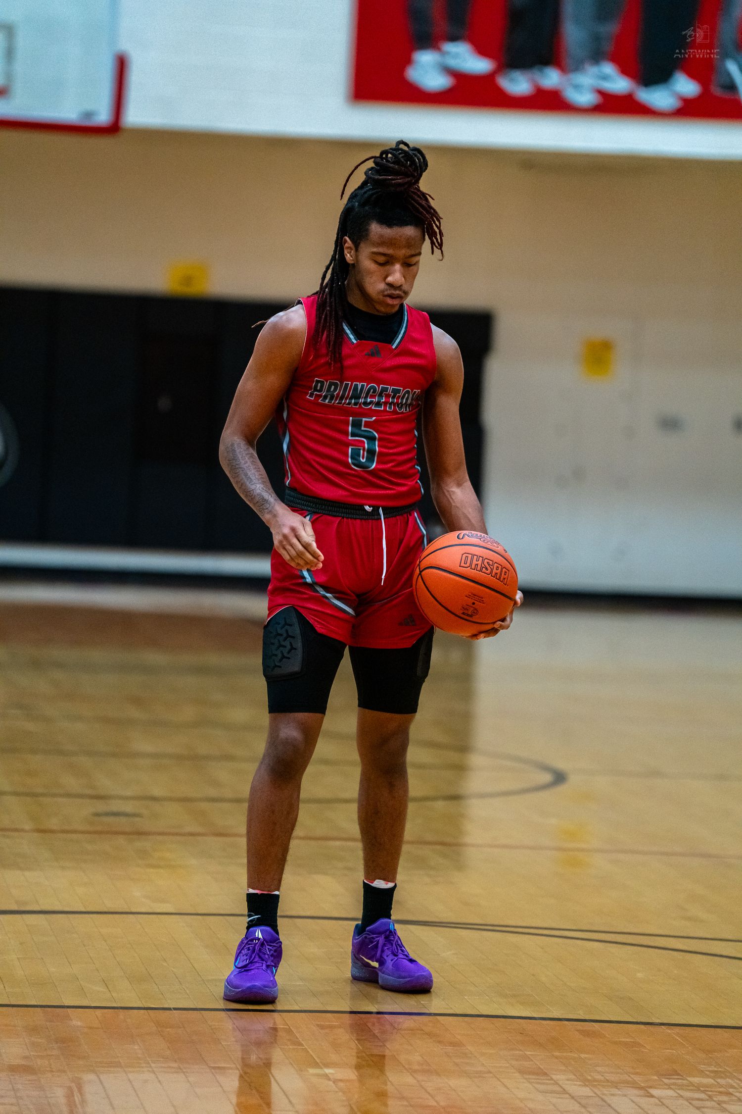 Princeton #5 player holds basketball while looking down during high school basketball game in red uniform.