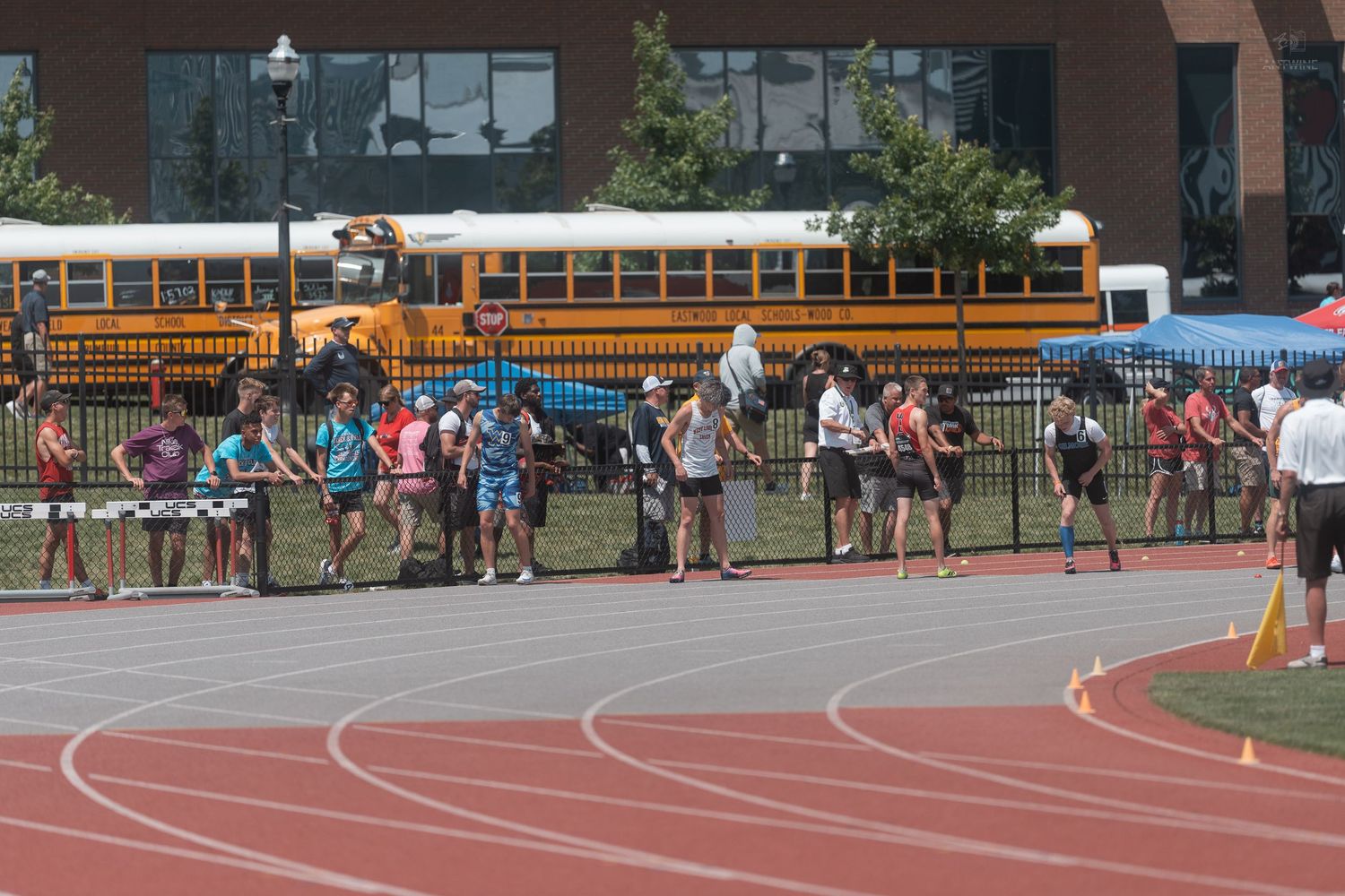 Students and spectators line up along a fence at an outdoor track and field event near yellow school buses.