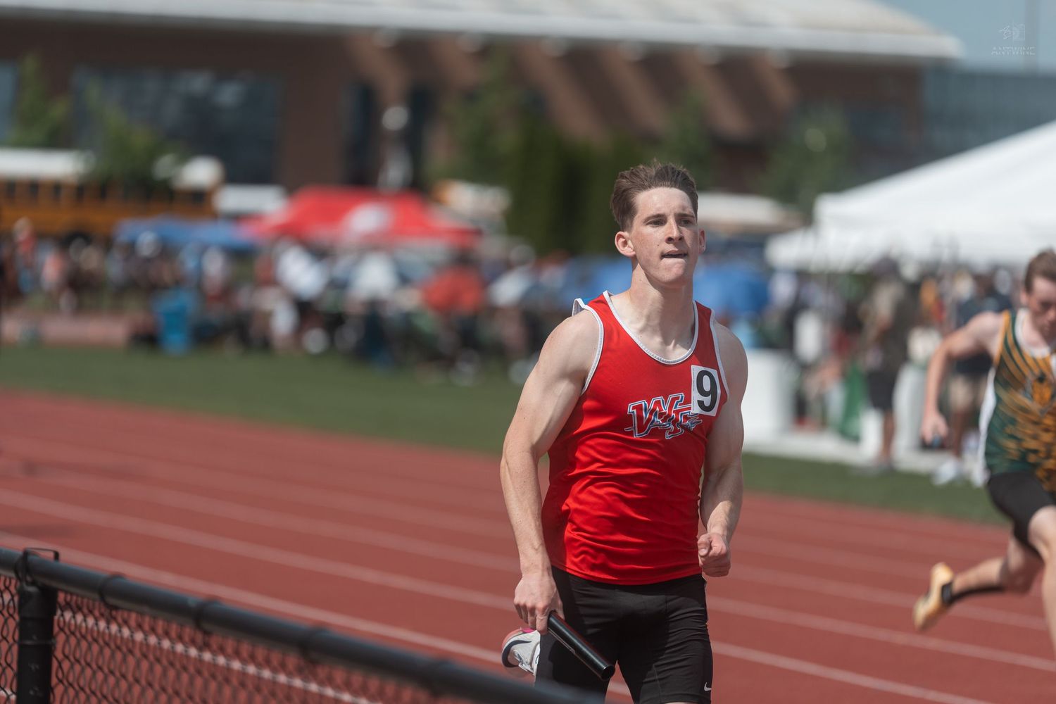 Male track athlete wearing number 9 red jersey runs competitively on outdoor track during race.
