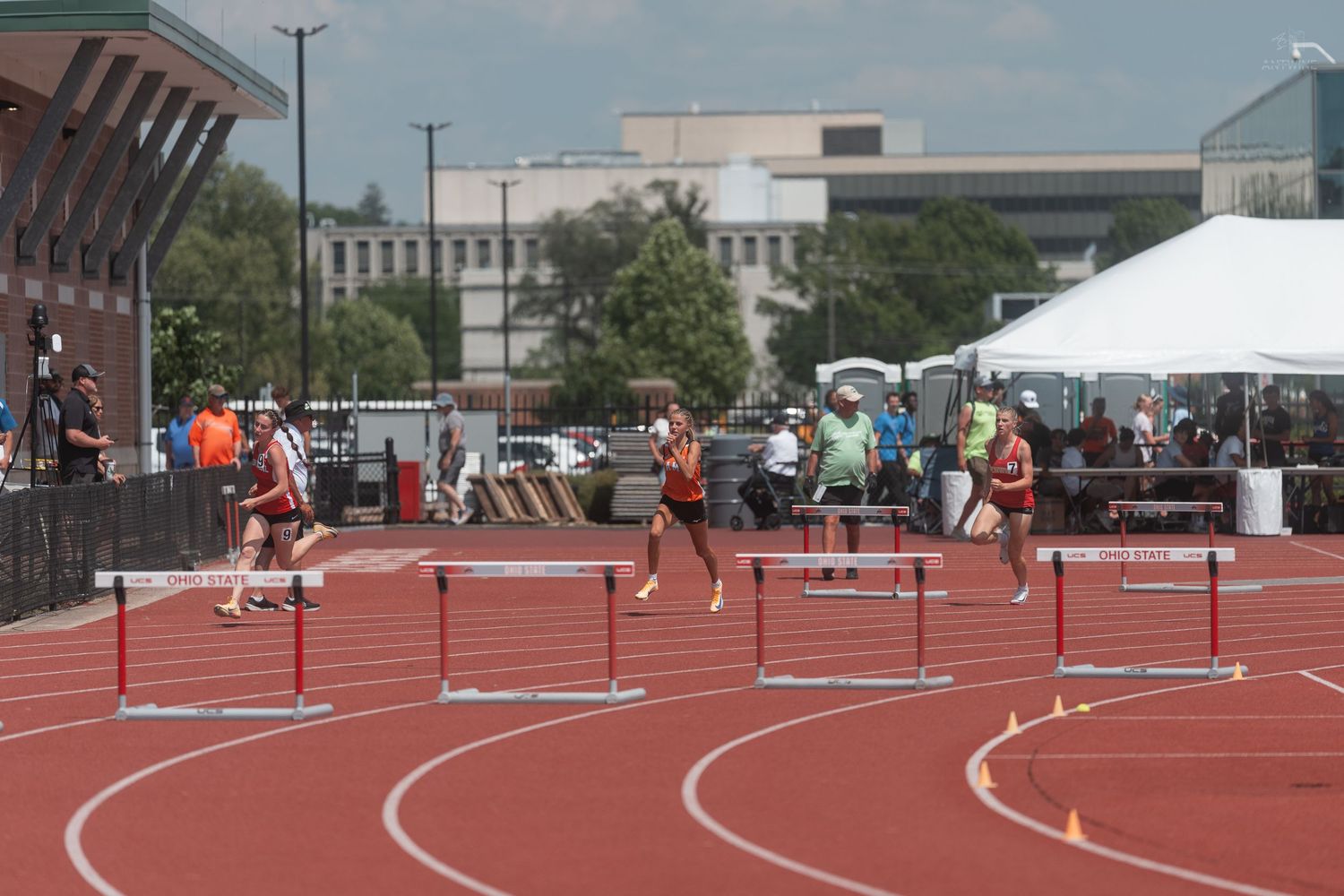 Athletes clearing hurdles on a red outdoor track during a competitive track and field meet under blue skies.