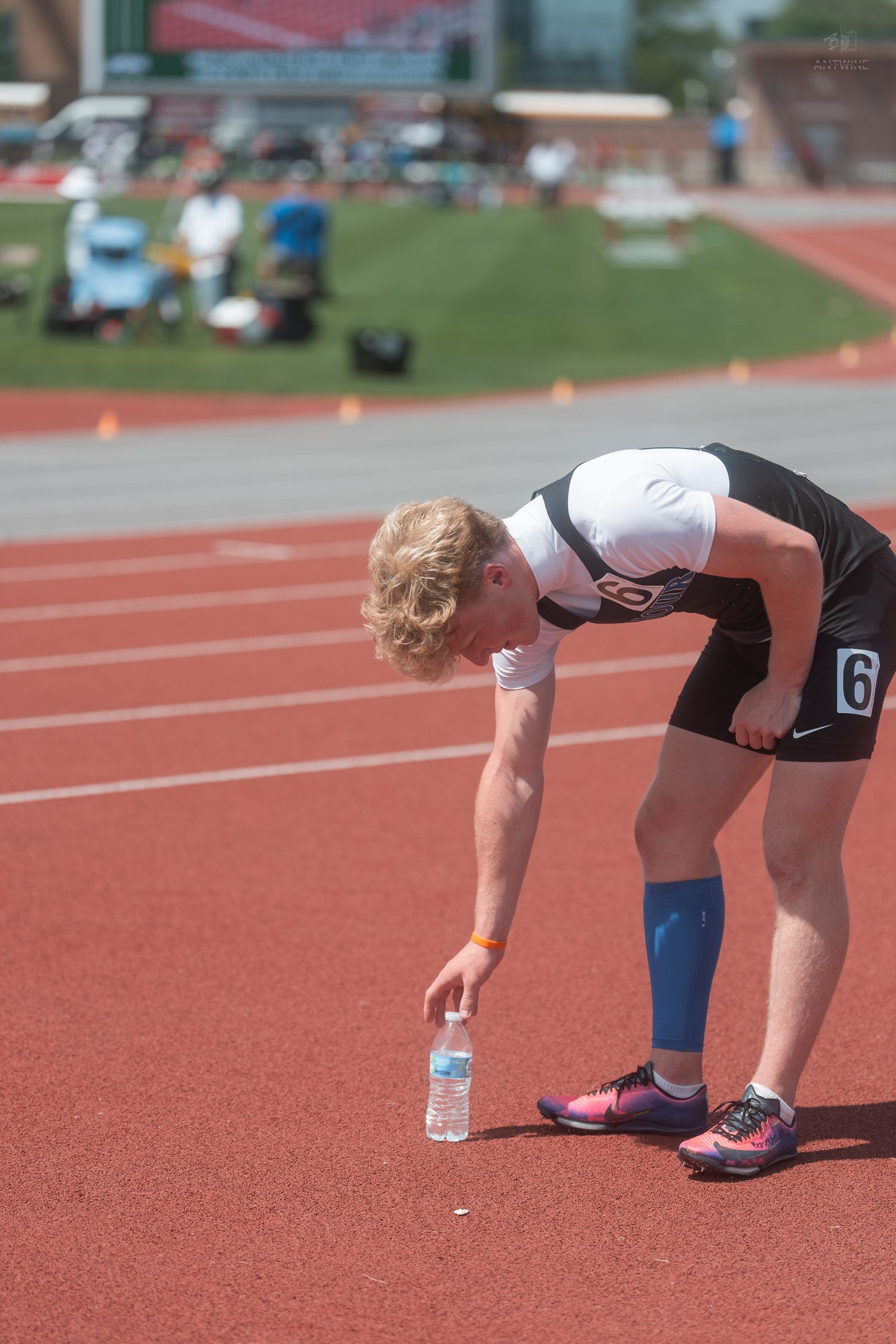 Young male track athlete number 6 bending down to pick up water bottle on red track during outdoor meet.