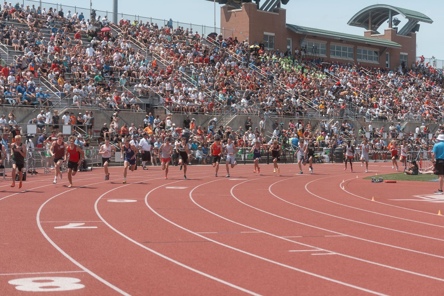 Runners compete on a red track during a packed high school track and field meet with a large crowd filling the stadium.