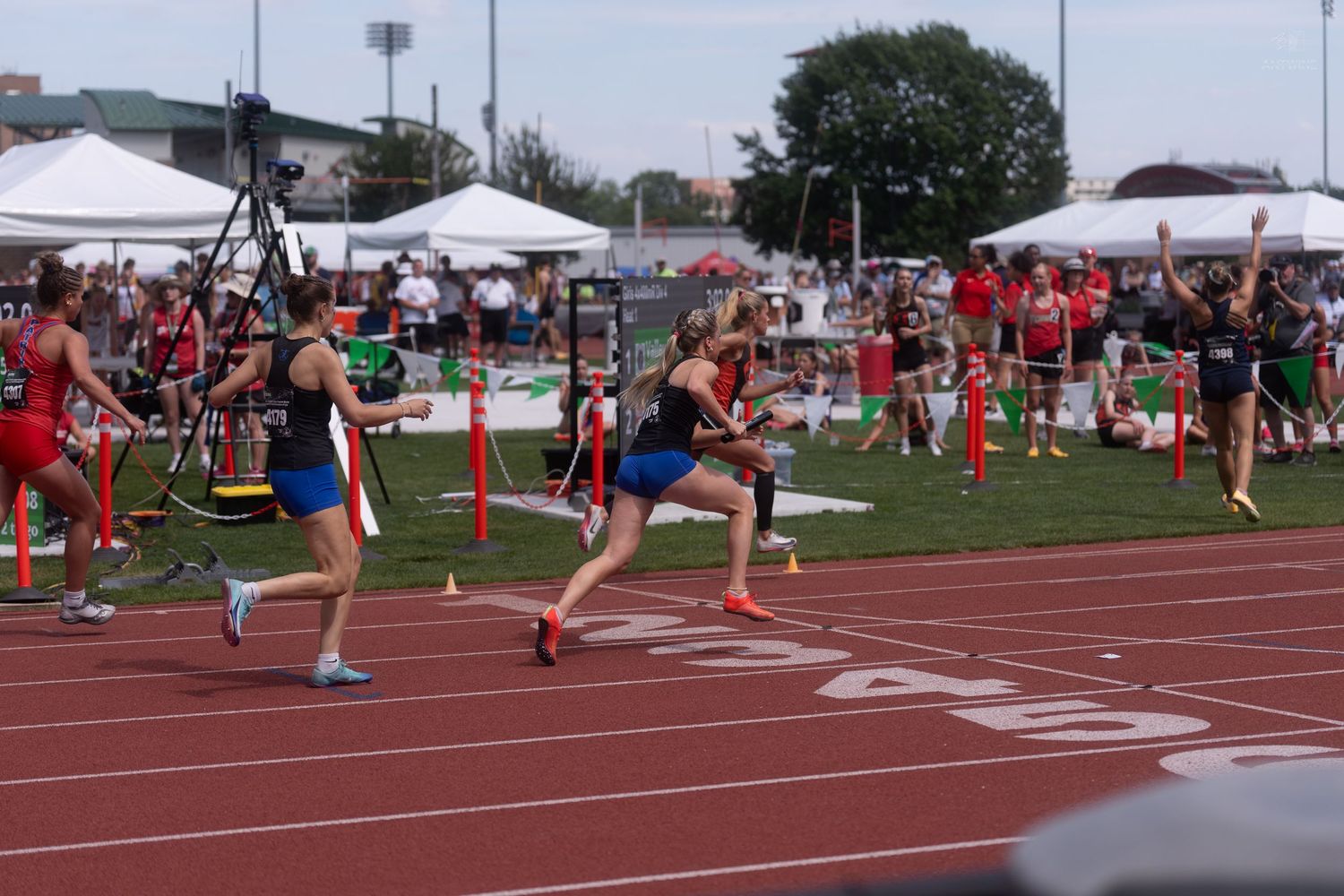 Runners compete in a sprint race at an outdoor track and field meet with spectators in the background.