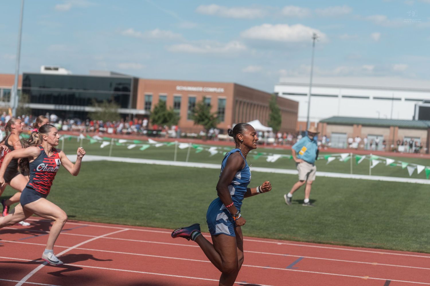 Female athletes compete in a sprint race on a red track during an outdoor track and field meet on a sunny day.