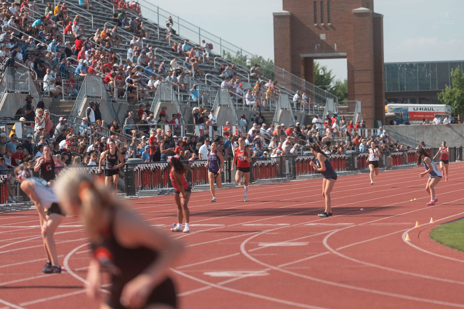 Athletes compete in a relay race on a red track as a large crowd watches from the stands.