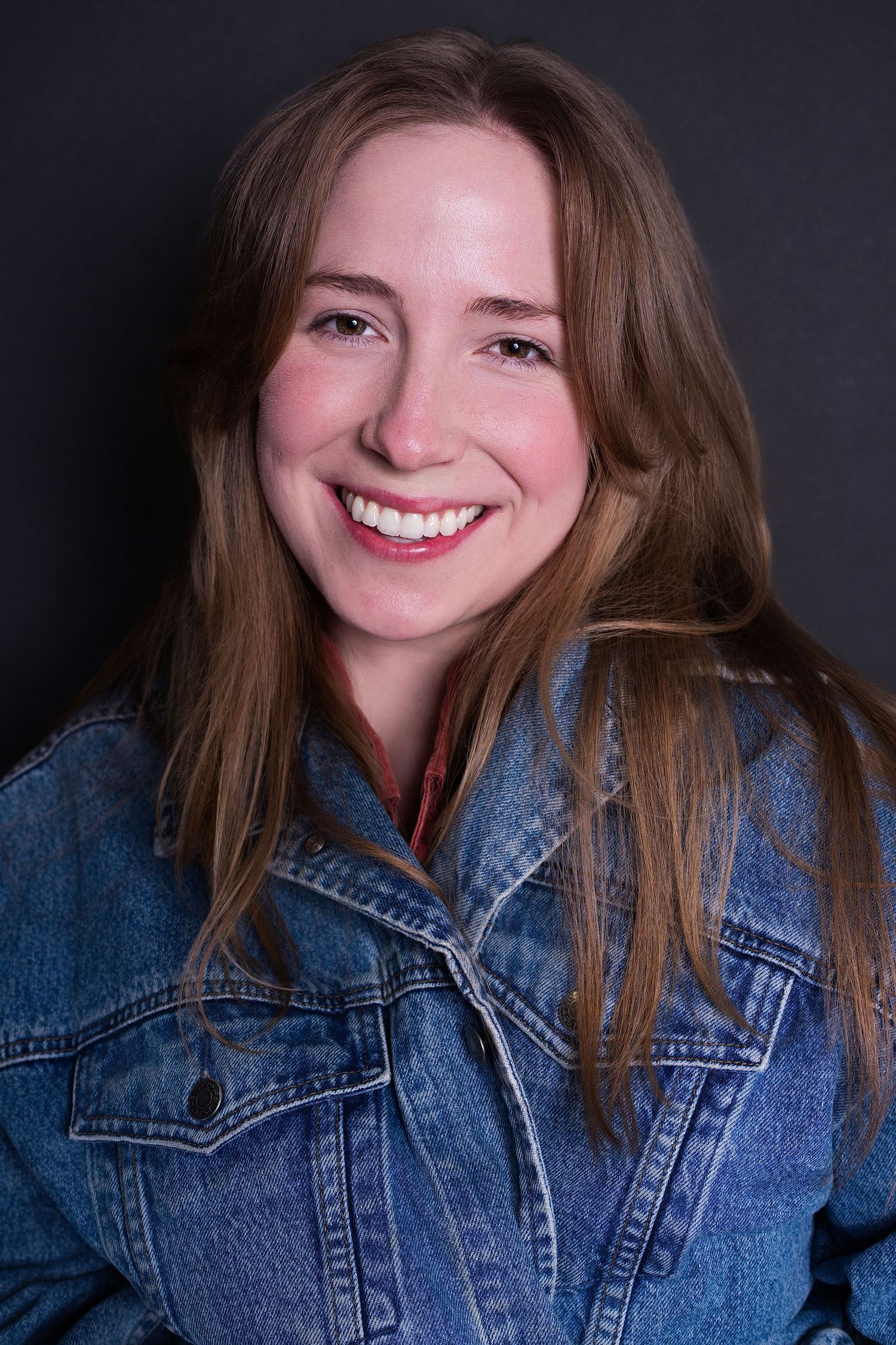 Cheerful young woman with long hair in blue denim jacket smiling brightly against dark gray background.