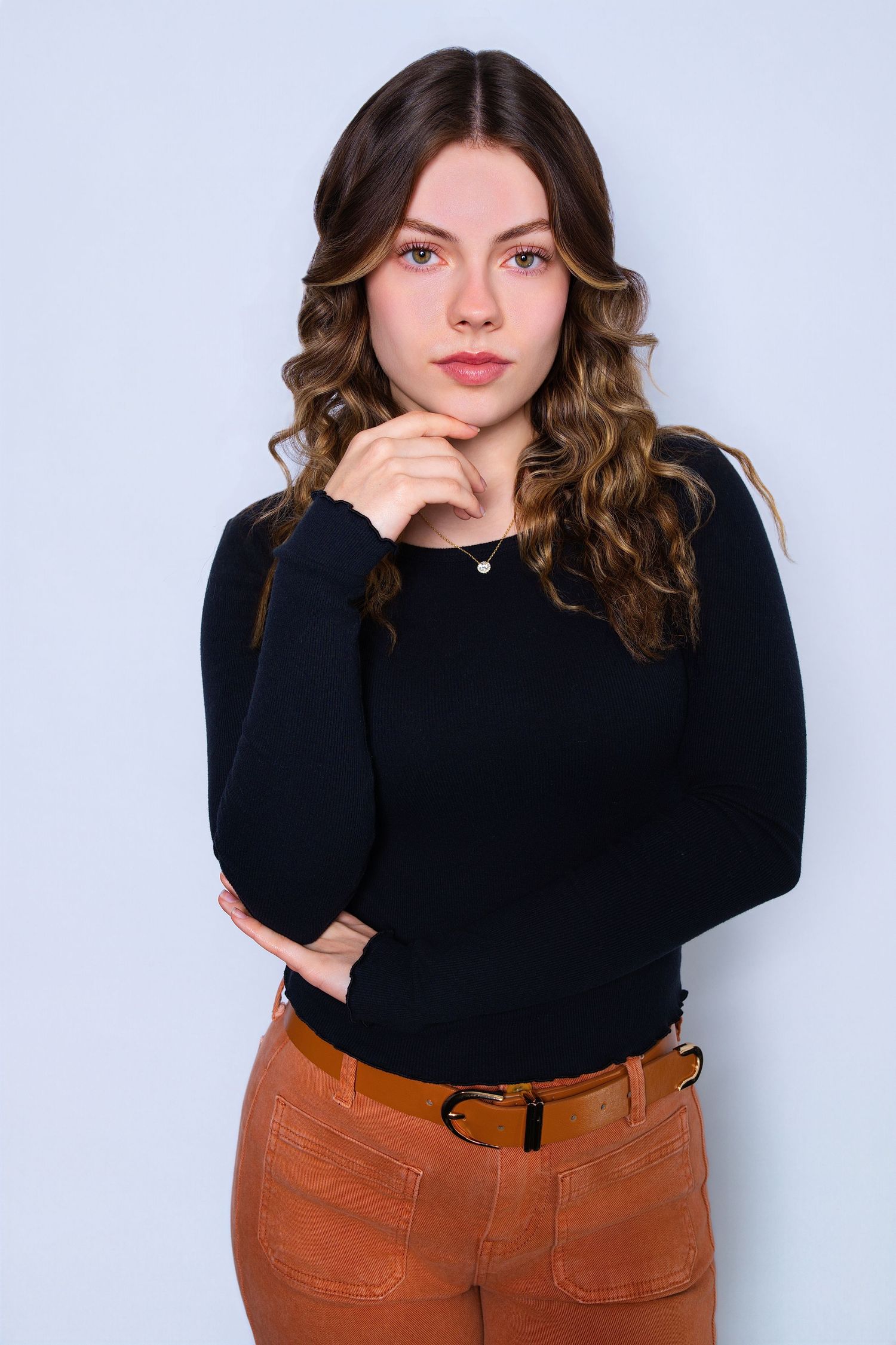 Young woman with curly hair in black sweater and brown pants poses thoughtfully.