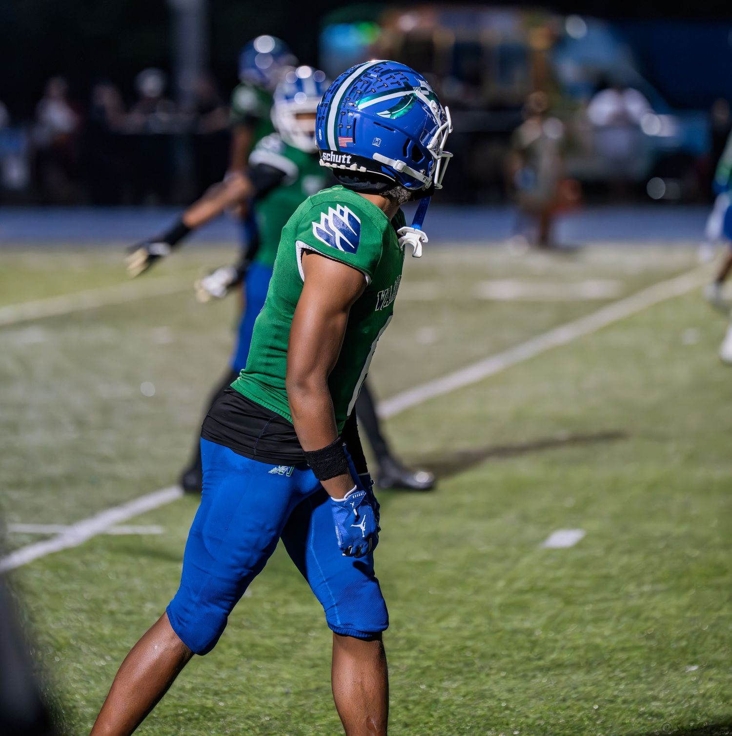 A football player in green and blue uniform walks on the field during a night game.