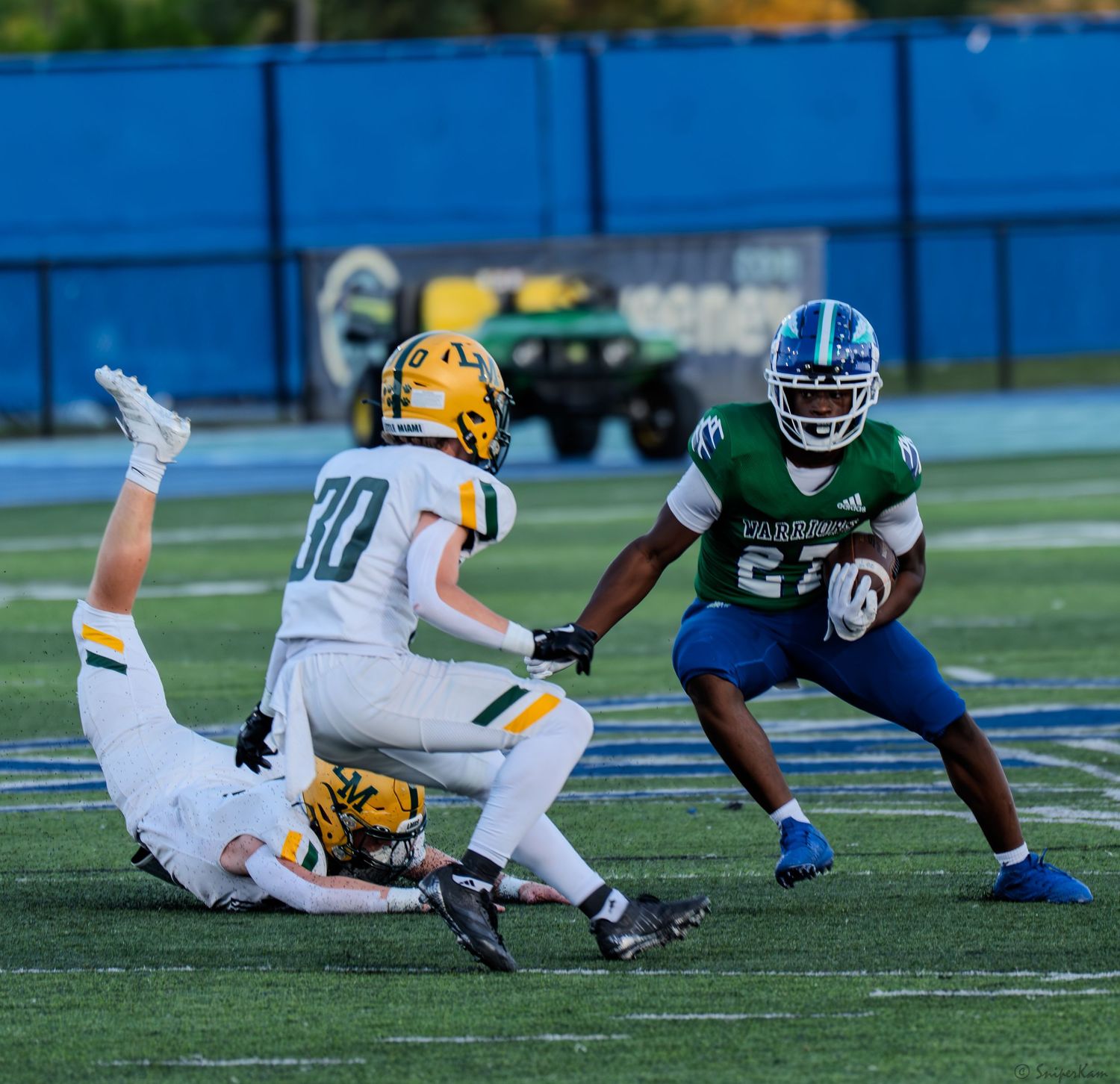 Football player in green uniform evades two defenders in white and gold uniforms on a blue turf field.