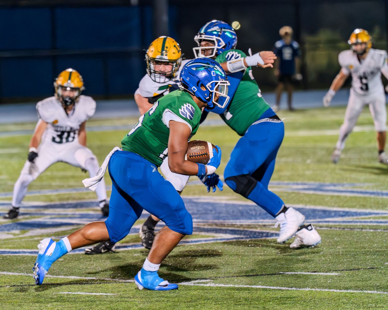 Warriors quarterback hands off to running back during a high school football game at night.