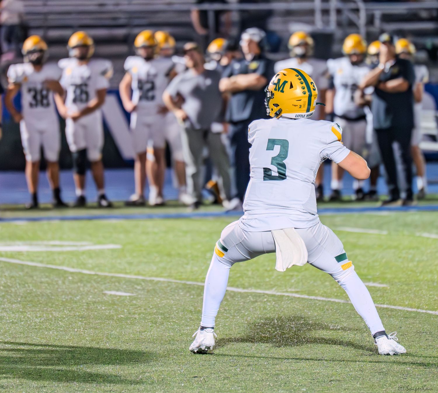 High school quarterback number 3 in white and gold prepares to throw during night football game on turf field.
