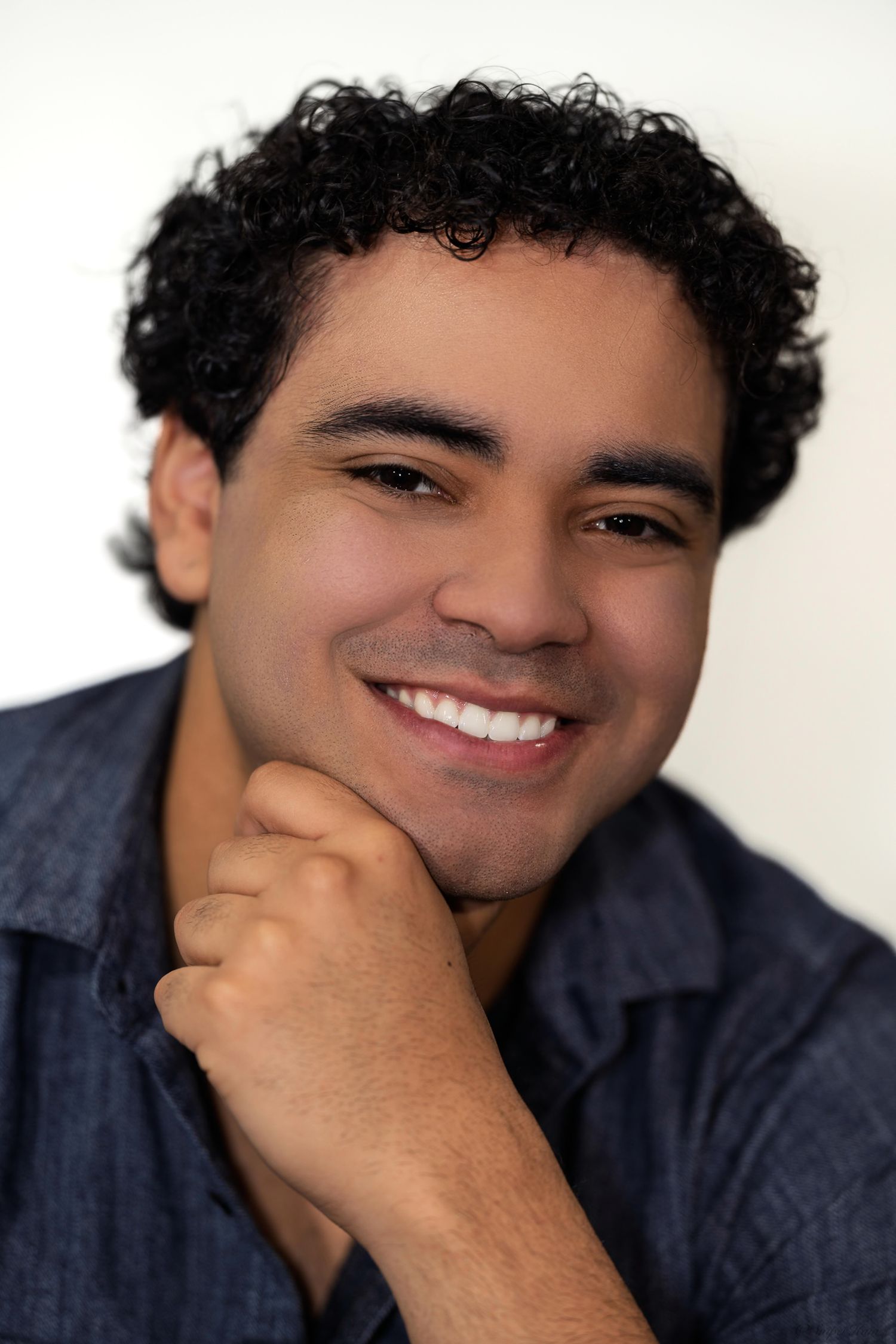 Smiling young man with curly black hair resting chin on fist against white background.