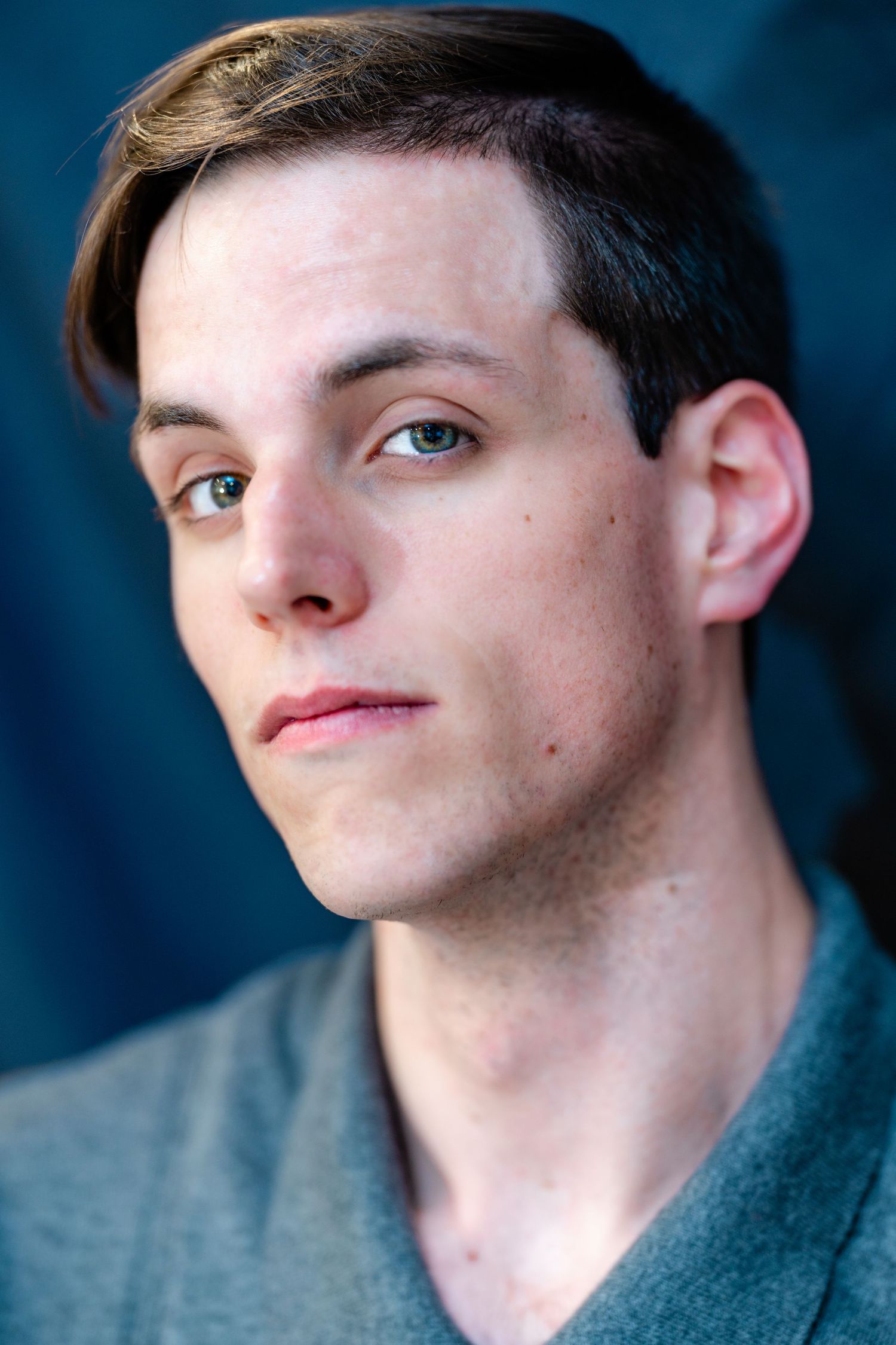 Young man with swept brown hair and light eyes photographed against dark blue backdrop.