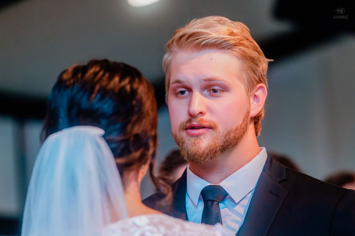 Groom with blonde hair and beard gazes lovingly at veiled bride during wedding ceremony.