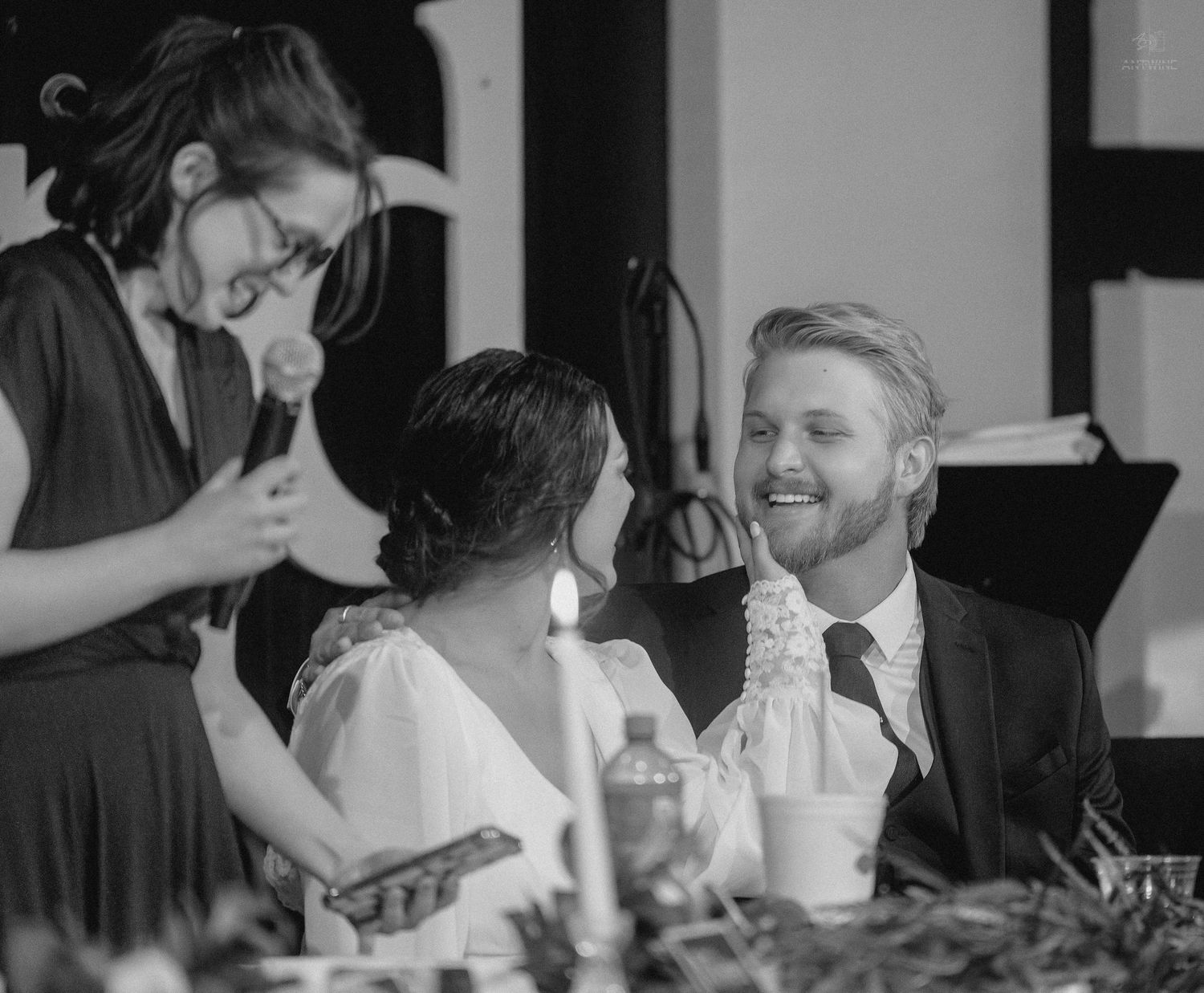Wedding guest gives speech to laughing bride and groom at candlelit reception dinner table.