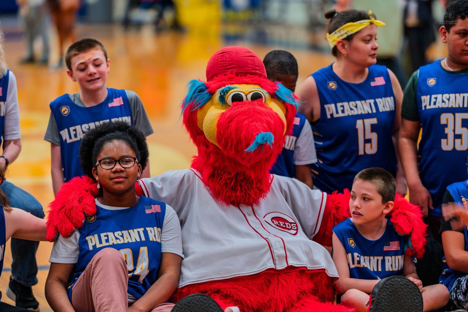 Red mascot poses with kids in Pleasant Run basketball jerseys at a gym event.
