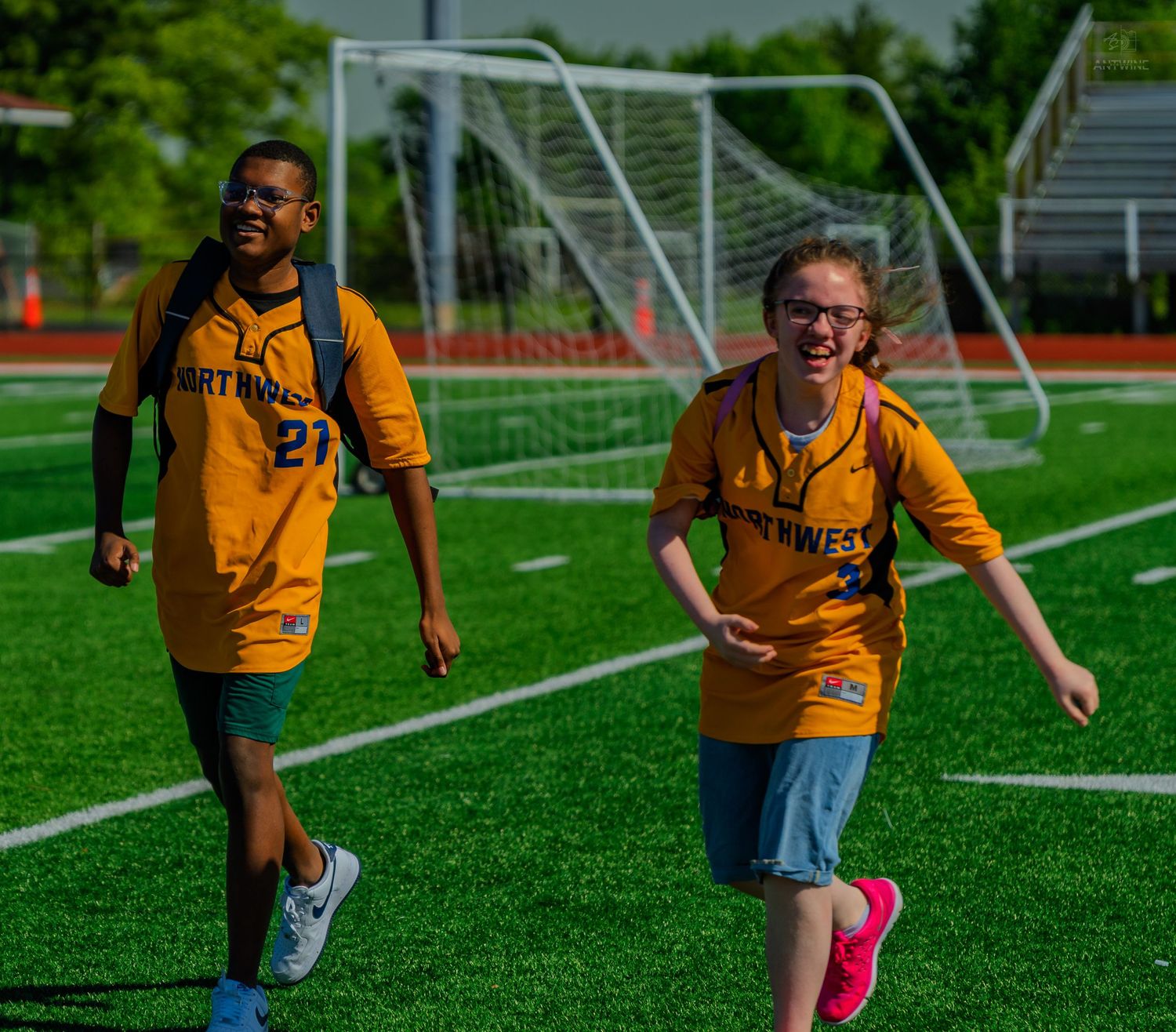 Two students in gold Northwest jerseys smile and run on a green turf soccer field during a school sports event.