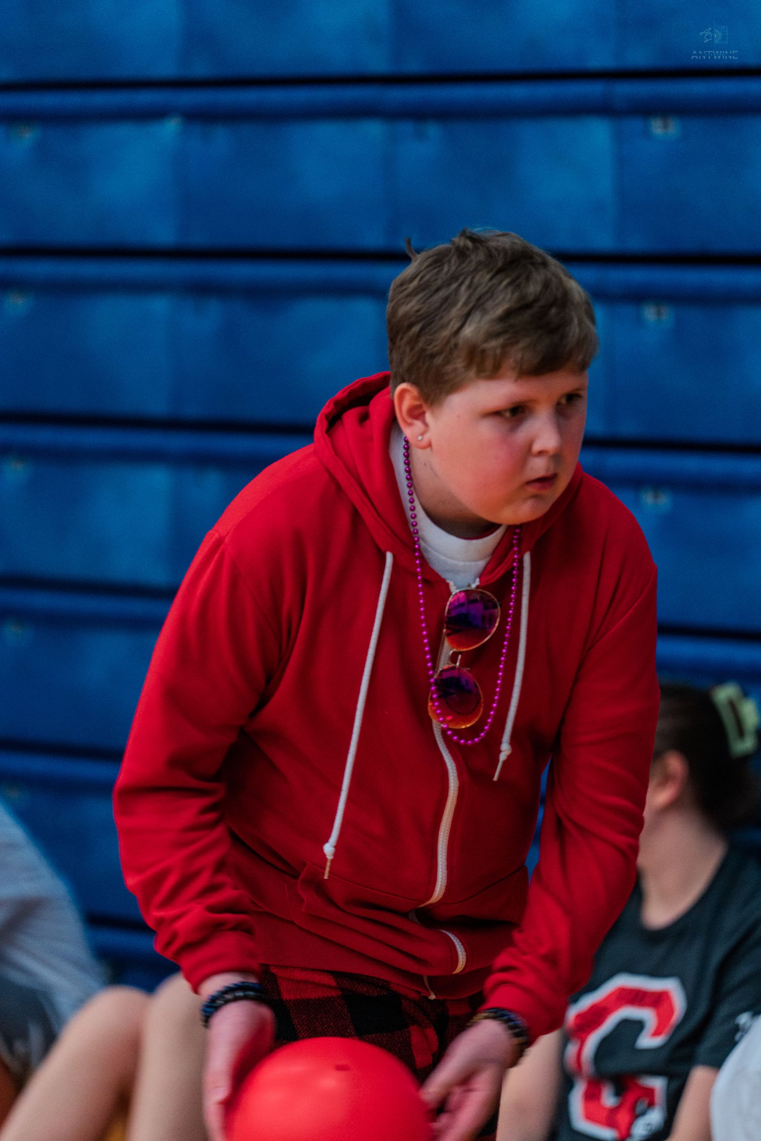 A young boy in a red hoodie wearing colorful beaded necklaces and sunglasses sits in blue bleachers.