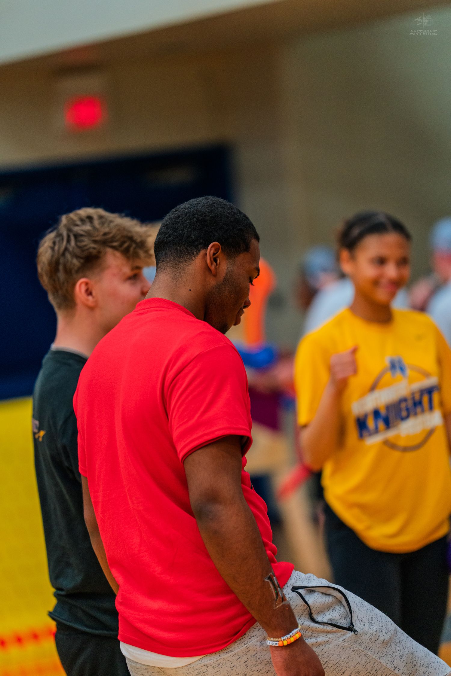 A man in a red t-shirt stands in a gym with teens, one wearing a yellow Knights jersey.