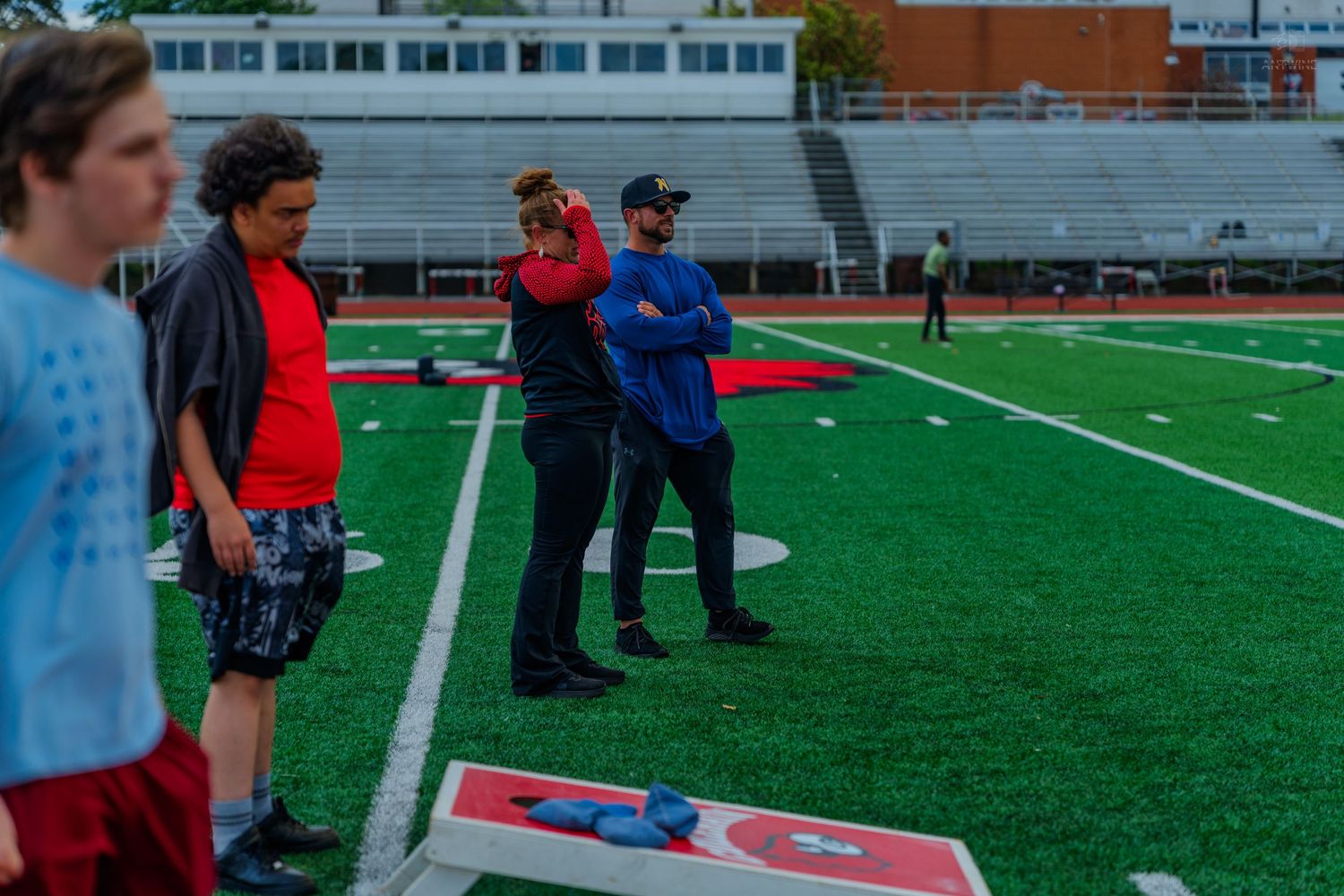 People playing cornhole on a green football field with stadium bleachers in the background.