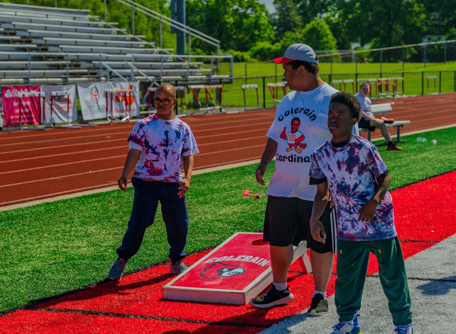 Kids and adult playing cornhole on red track at Cardinal sports event on sunny day.