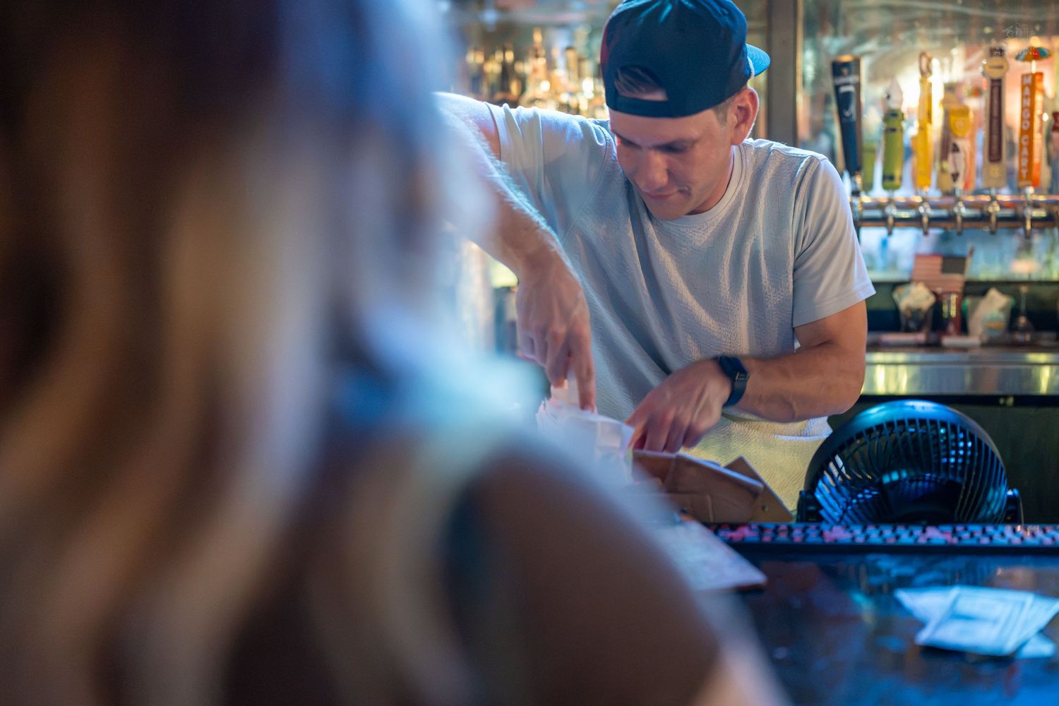 Bartender in white t-shirt and cap points at menu for customer at bar counter.