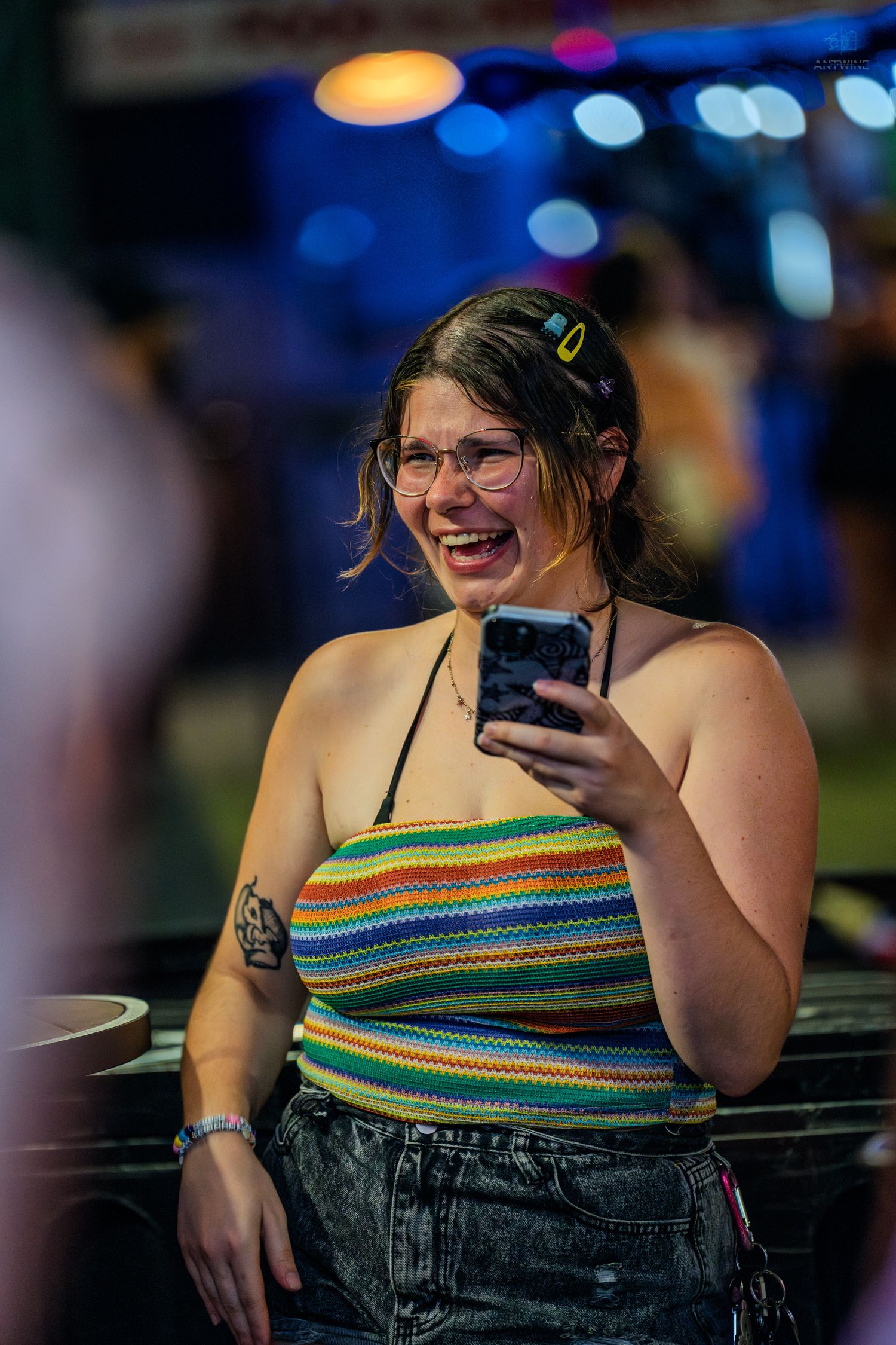 A smiling young woman with glasses holds a phone at a colorful nighttime outdoor event wearing a rainbow striped crop top.