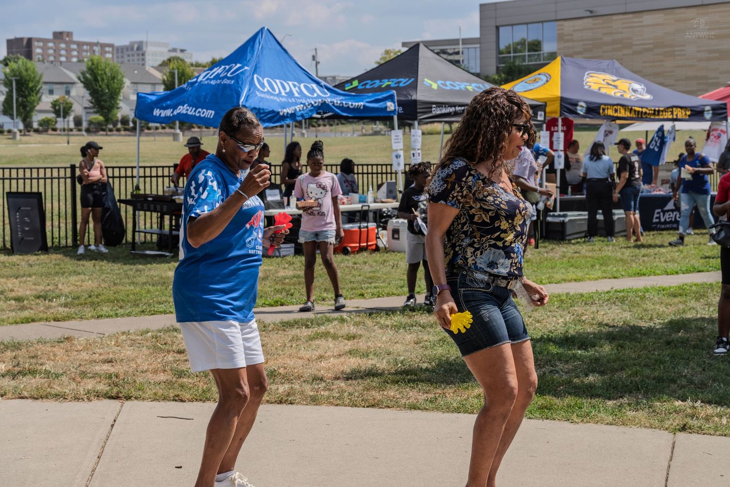 Two women dancing outdoors at a summer community festival with colorful vendor tents in the background.