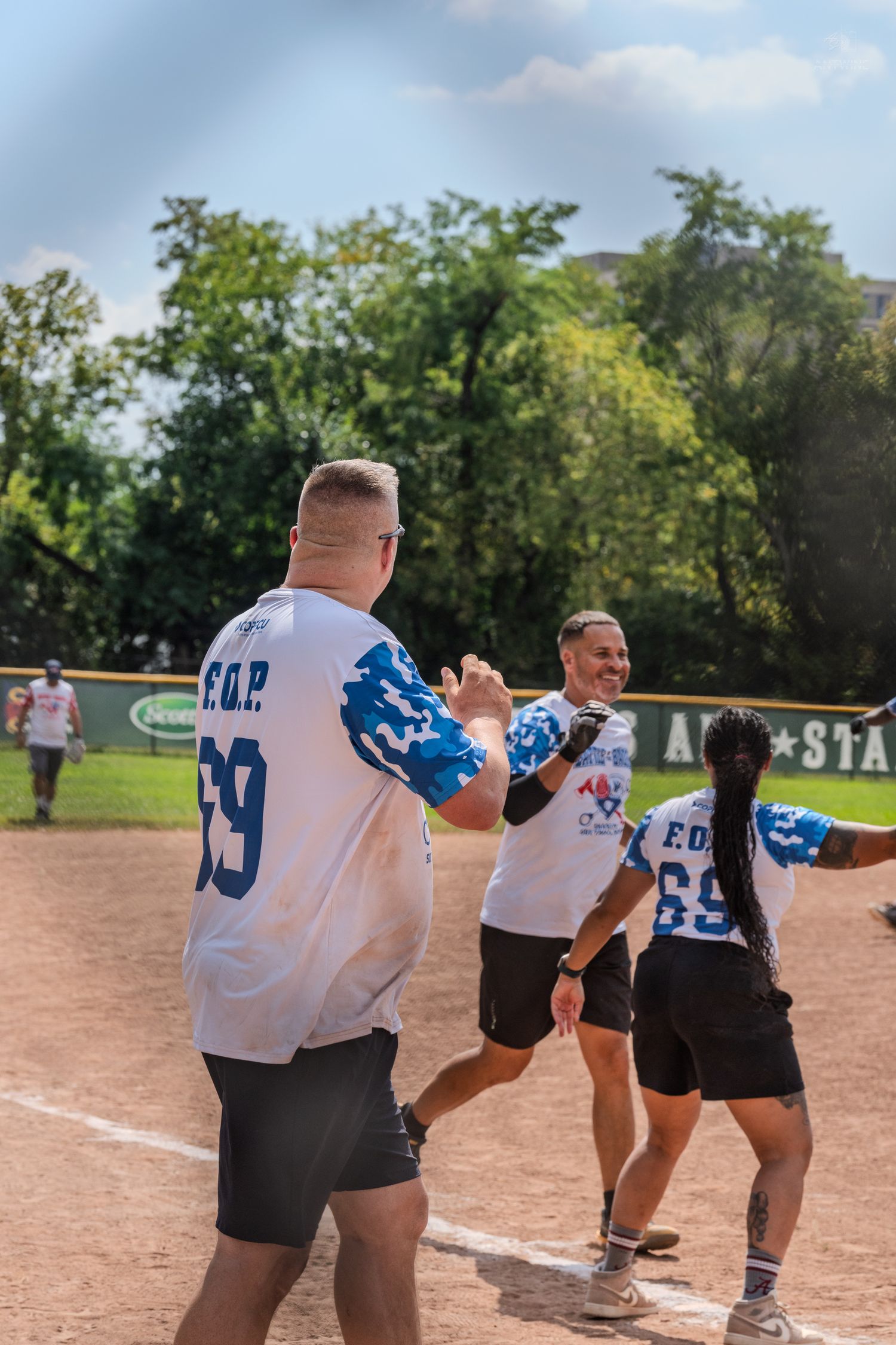 Players in EOP jerseys celebrate with high fives on a sunny softball diamond during a charity game.