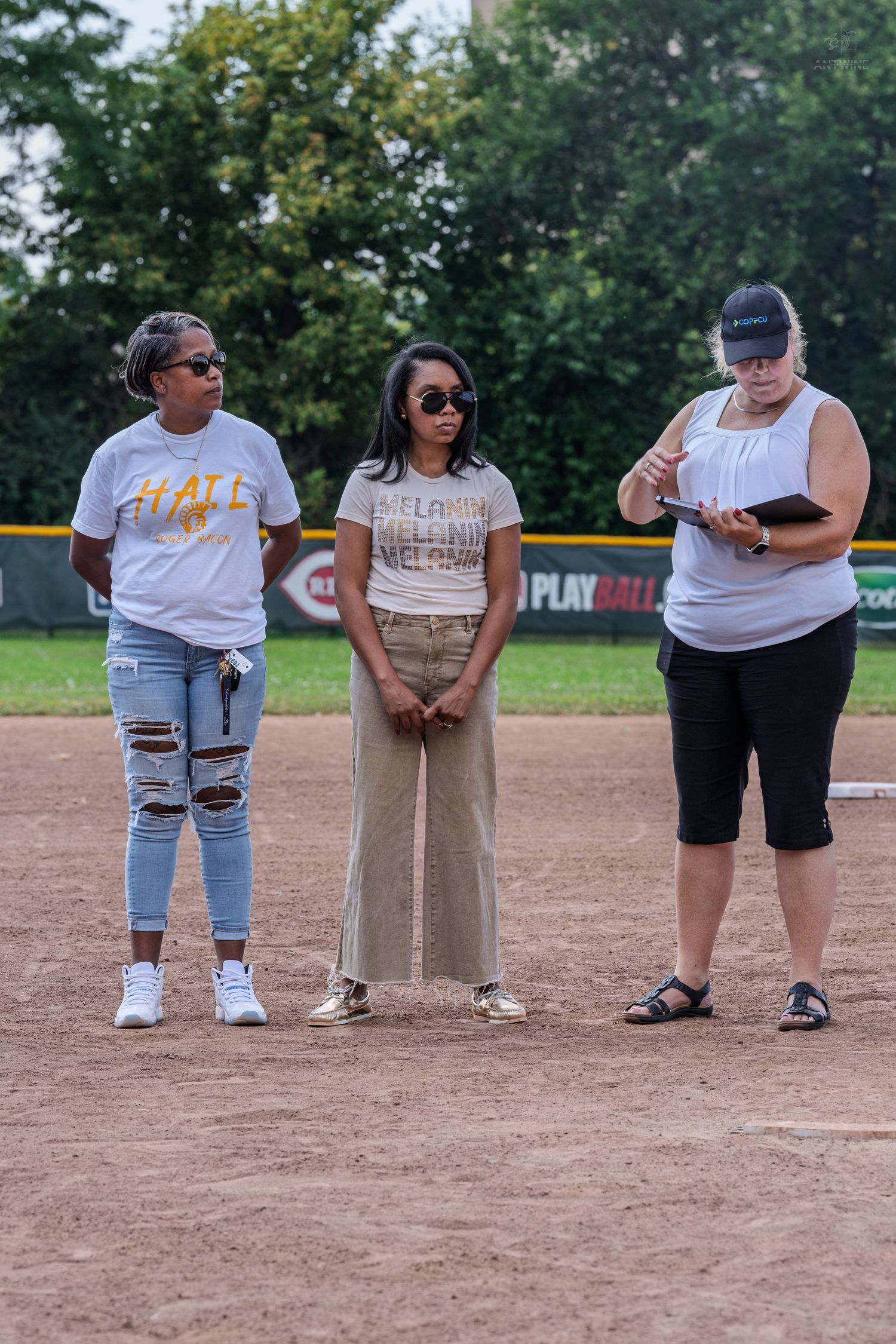 Three women stand on a baseball field dirt infield, two wearing graphic tees and one reading from a notebook.
