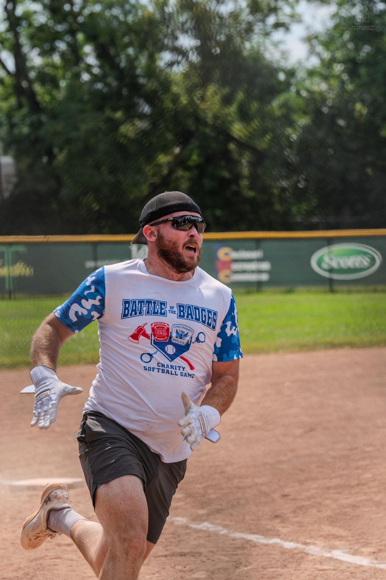 A man in a Battle of the Badges charity softball game shirt runs the bases during an outdoor softball game.