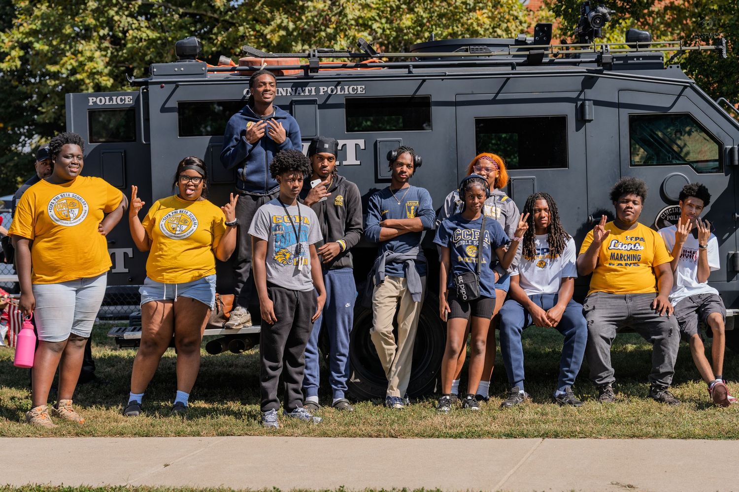 Group of youth and adults in gold shirts posing outdoors near a police vehicle on a sunny day.