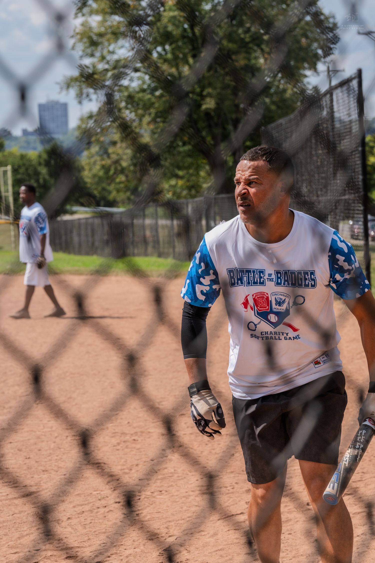 Male softball player holding bat wearing Battle of the Badges charity game jersey on field.