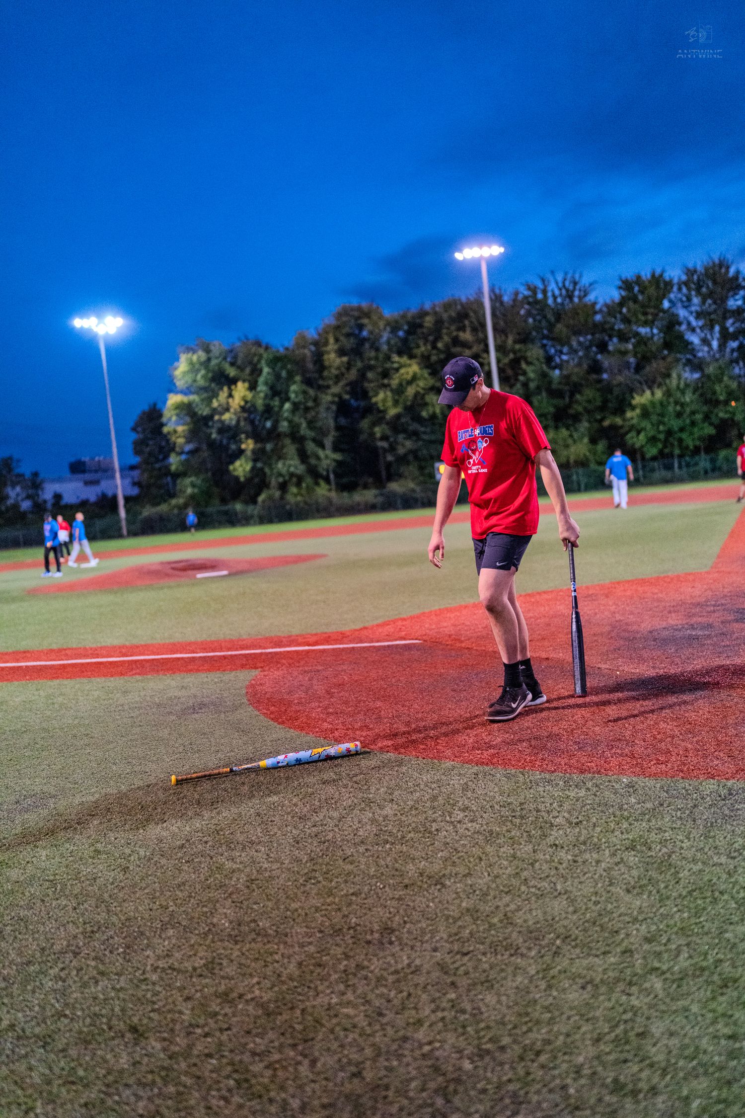 Softball player in red jersey stands near home plate holding a bat during a night game under bright field lights.
