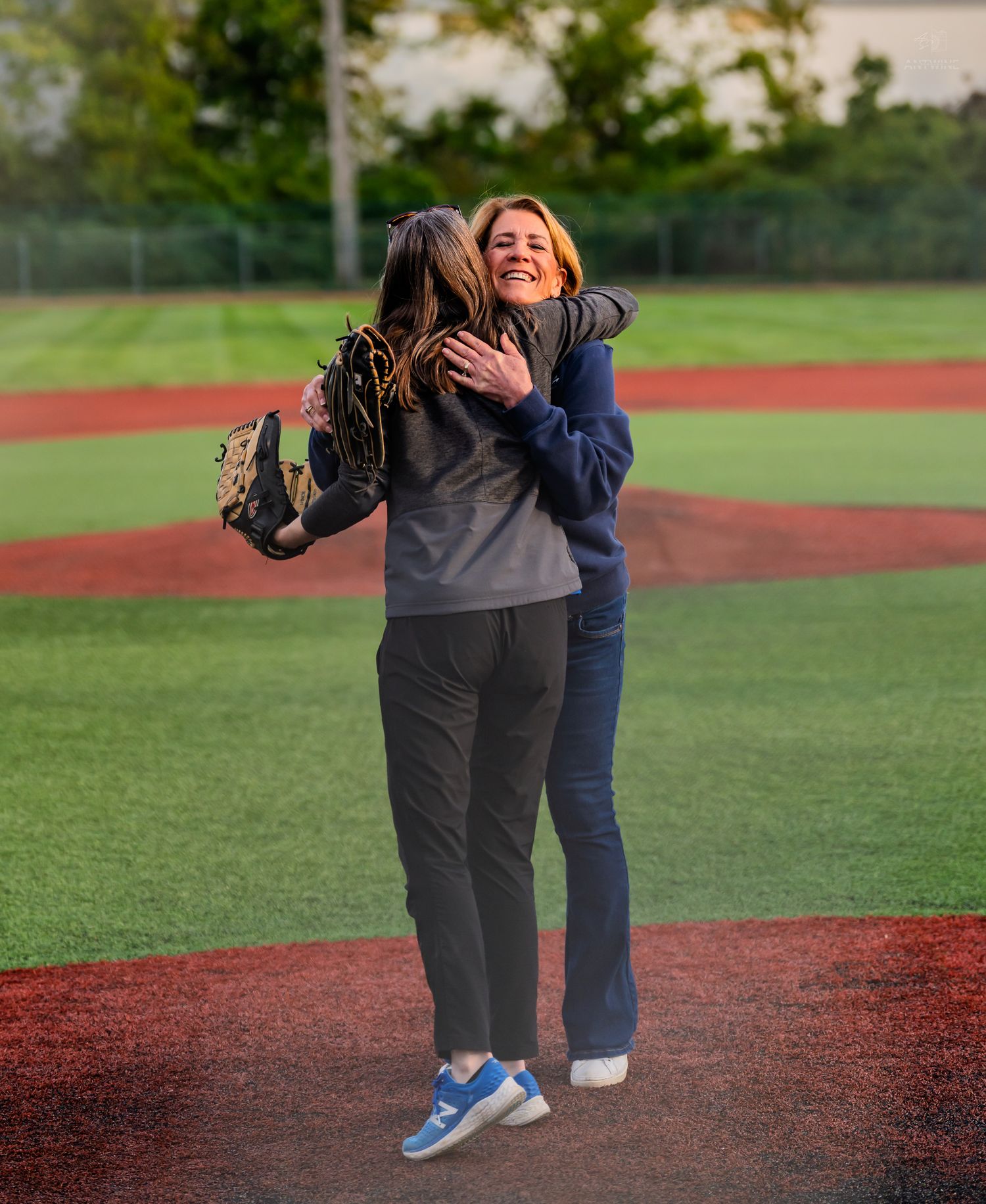 Two women share a joyful hug on a turf baseball field during a charity softball game.