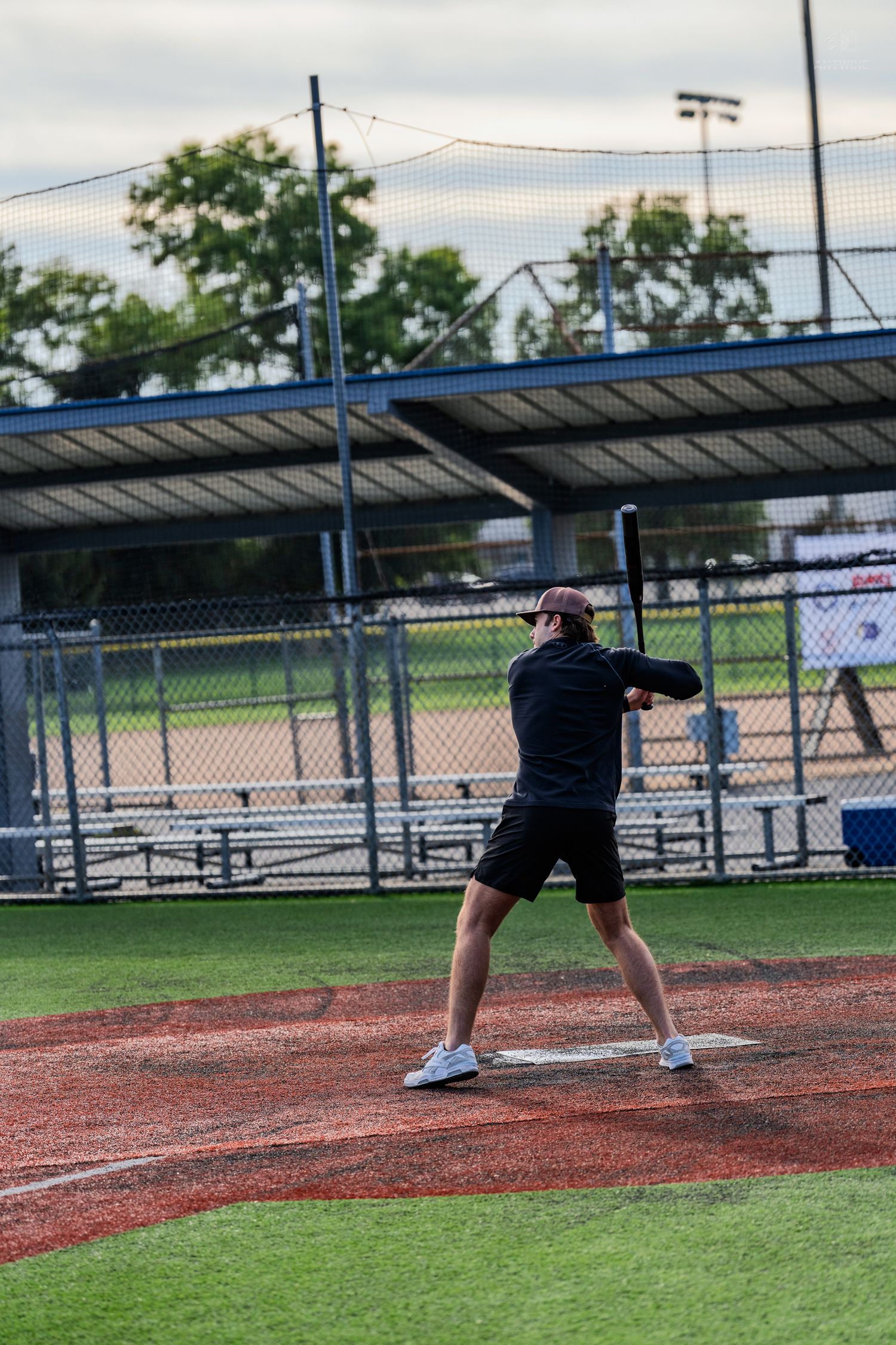 A baseball player in black athletic wear swings a bat on a red dirt field at an outdoor baseball diamond.