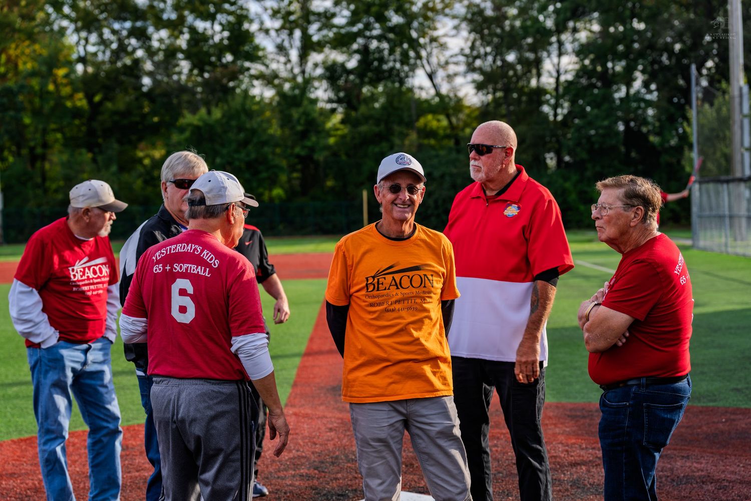 Adults in red and orange jerseys gathered on a baseball diamond during an outdoor softball event.