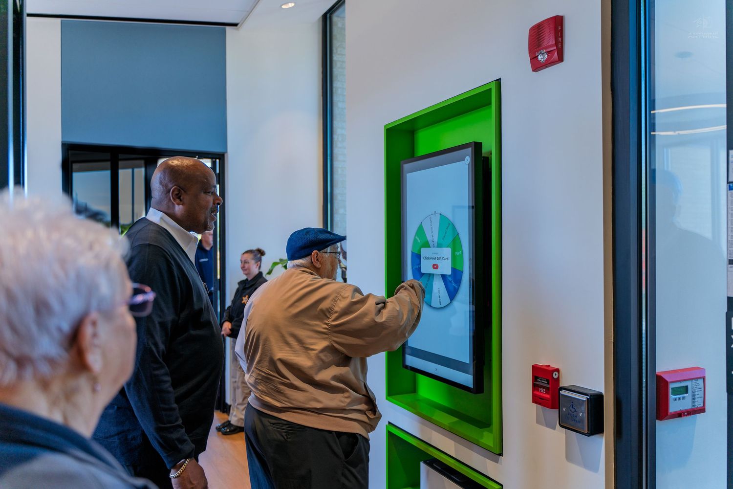Person using a green-framed interactive touchscreen kiosk in a modern building lobby with others waiting nearby.