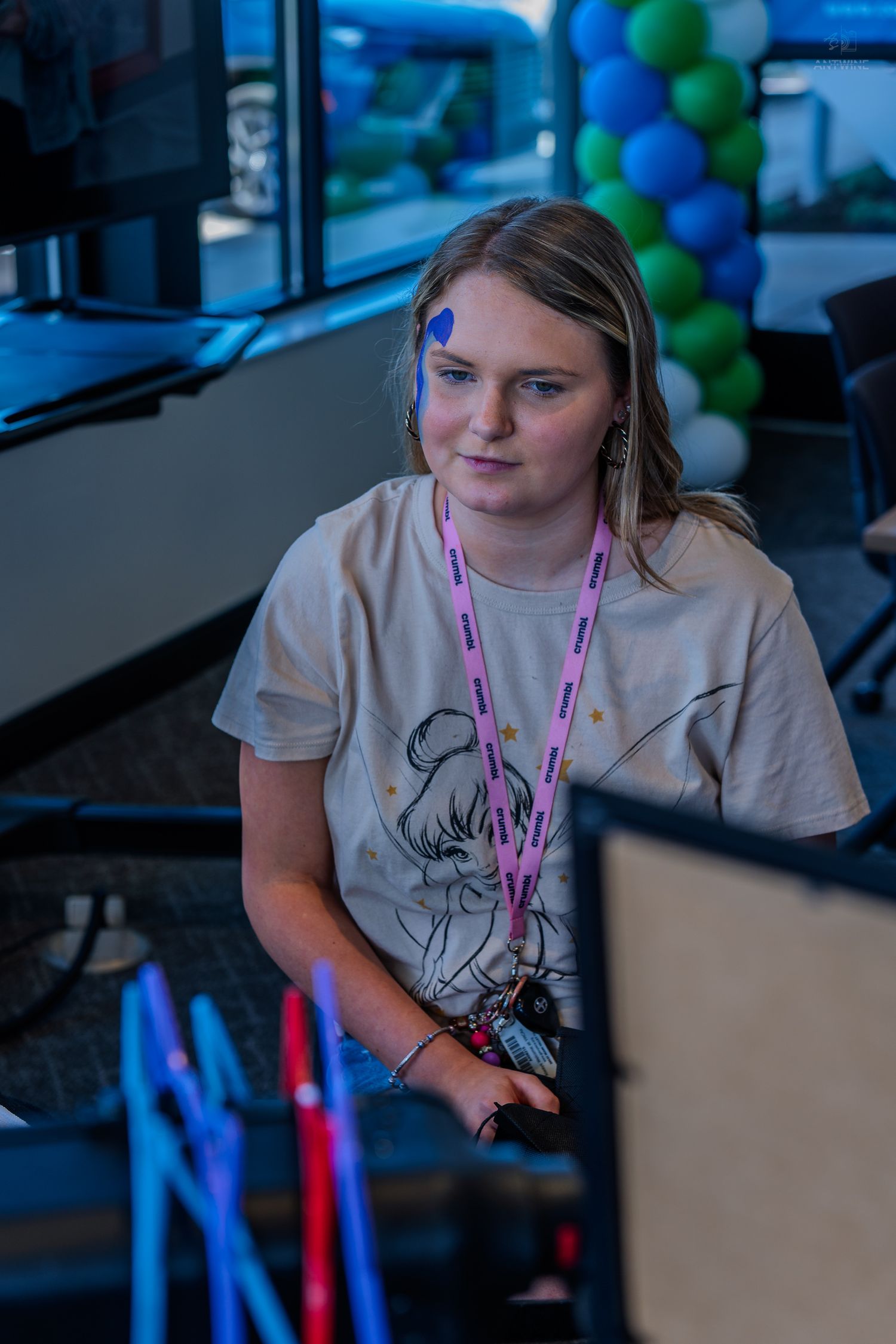 Young blonde woman with a blue dot on her face working at a computer during a festive event.