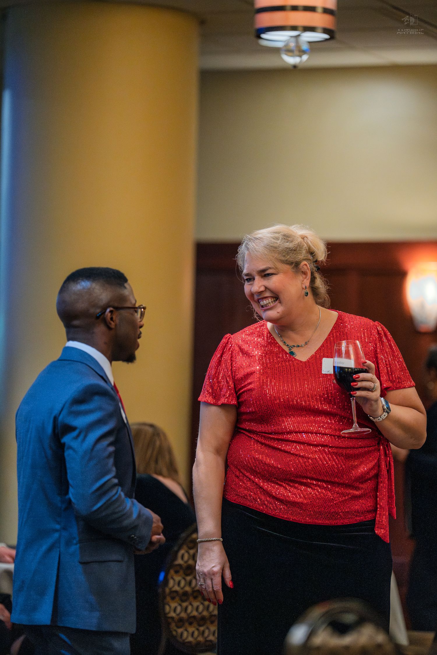 A woman in a red top holding a wine glass laughs while conversing with a man in a blue suit at a formal event.