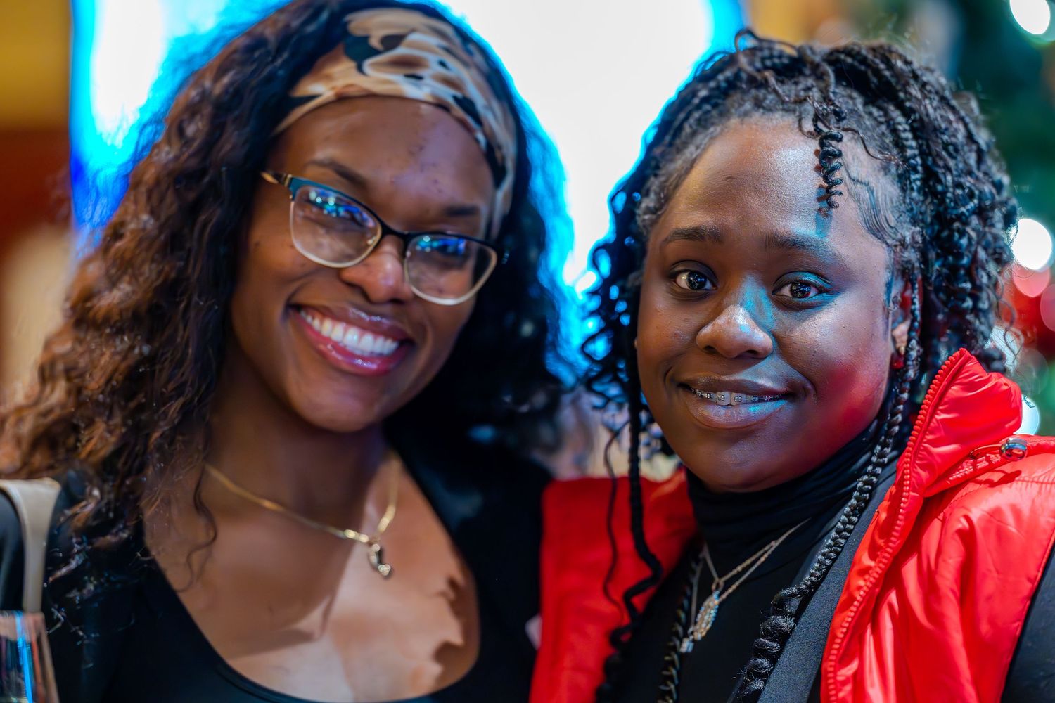 Two young women smiling together at an indoor event, one wearing glasses and a headband, the other in a red jacket.