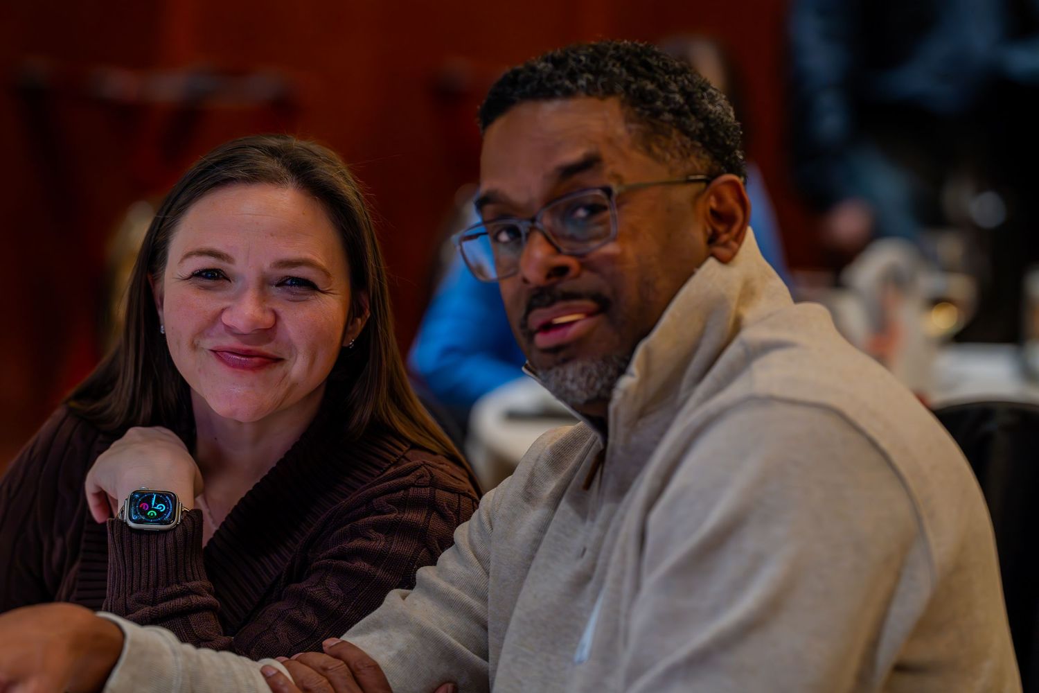 A smiling woman and man seated closely together at a table, sharing a warm moment at an indoor gathering.