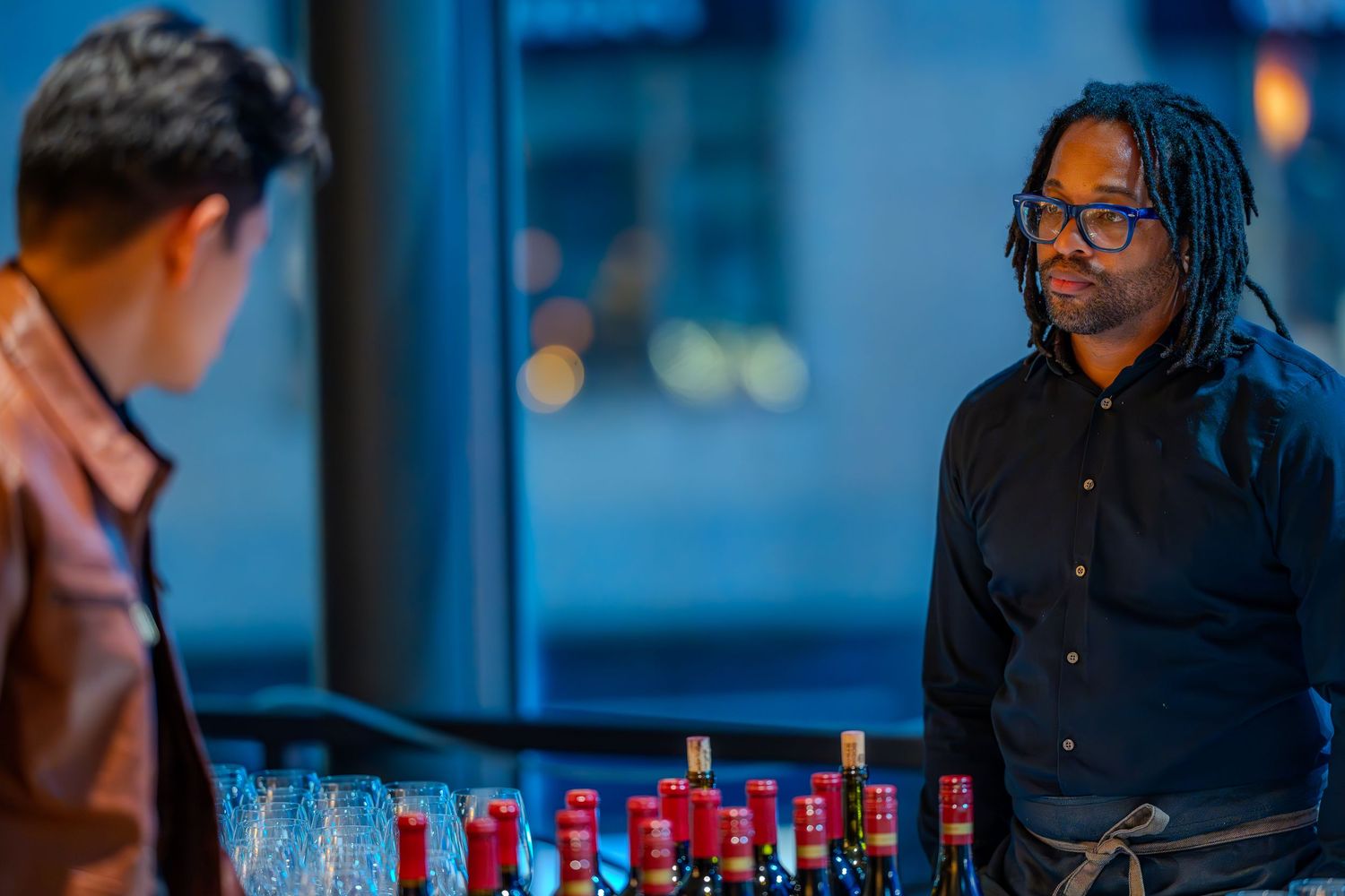 Bartender with dreadlocks in black shirt stands behind wine bottles at an upscale bar event.
