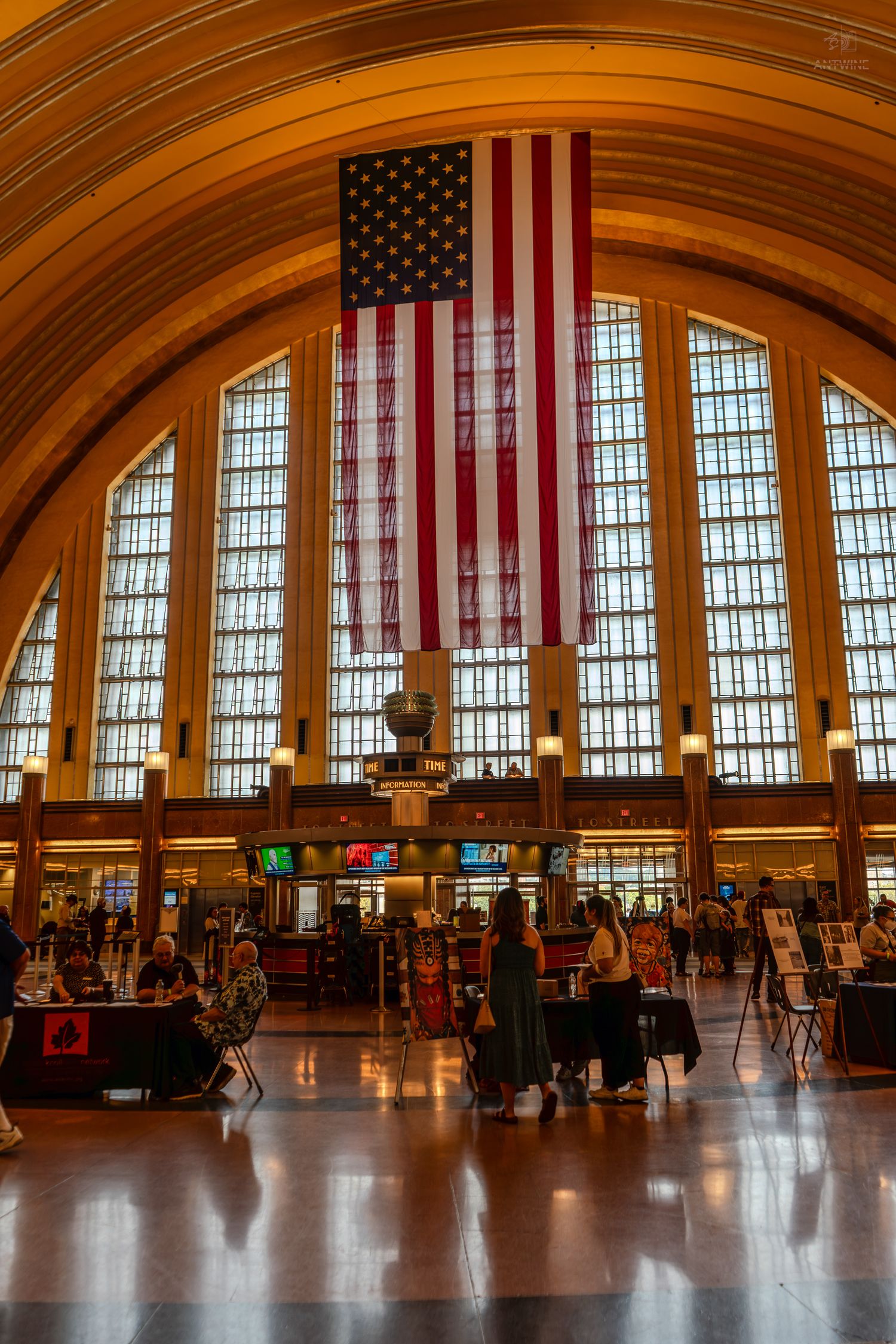 Grand hall of Cincinnati Union Terminal with large American flag hanging above arched windows and busy visitors below.